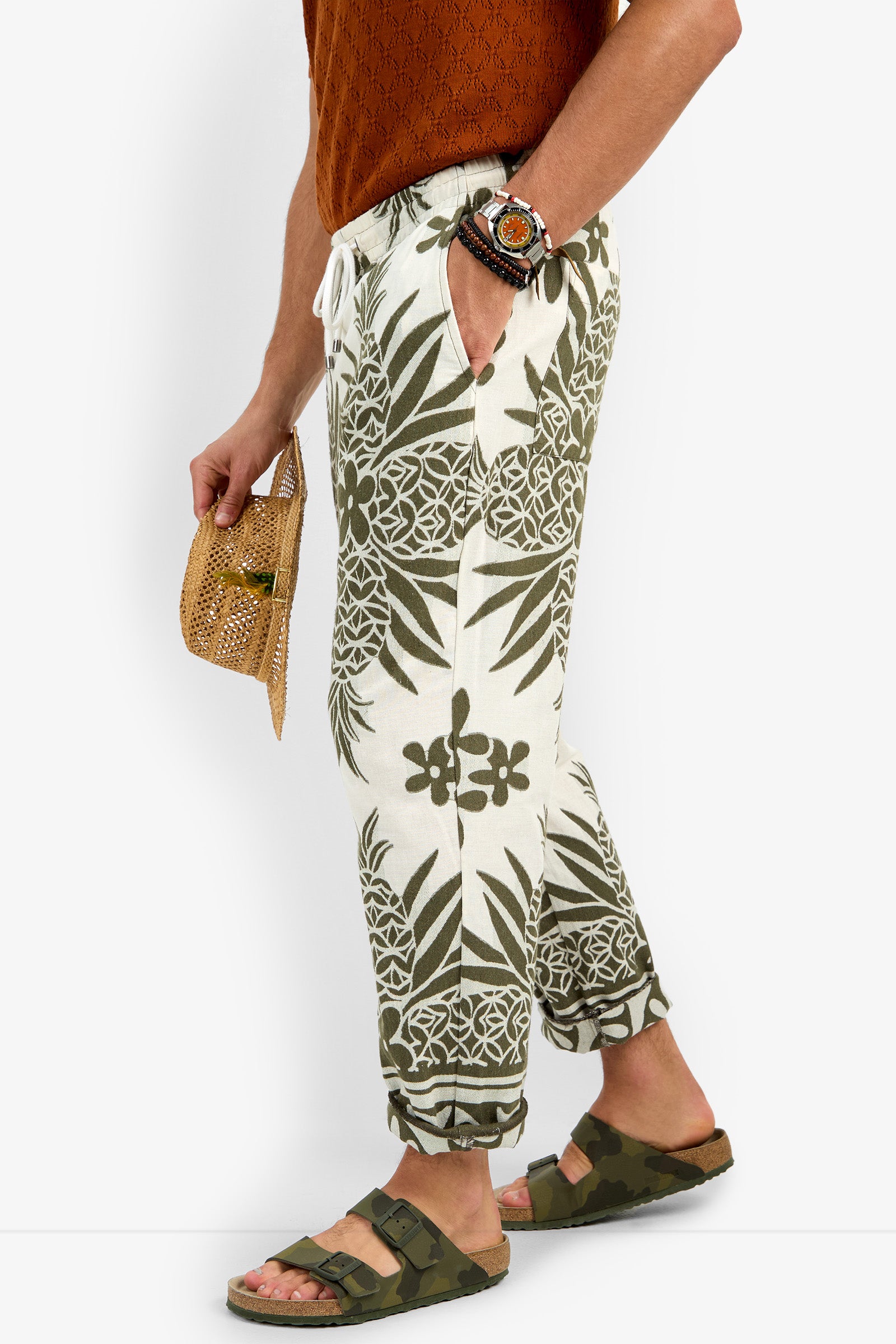 A person in Felipe Drawstring Pant with green tropical prints, an orange textured shirt, green sandals and bracelets, holding a small woven handbag and sunglasses.