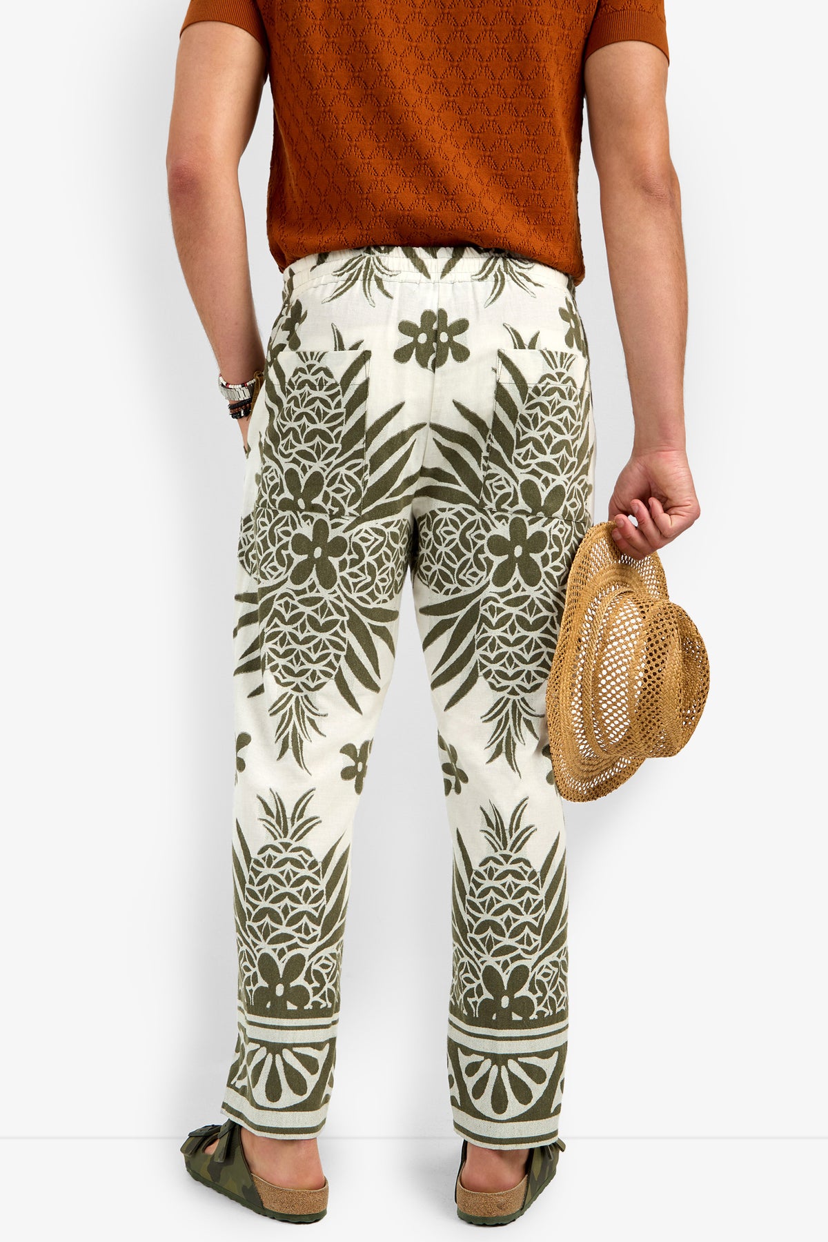 A person faces away, wearing a burnt orange short-sleeve shirt and Felipe Drawstring Pant in white with olive green pineapple and floral patterns, holding a woven straw hat and wearing green sandals.