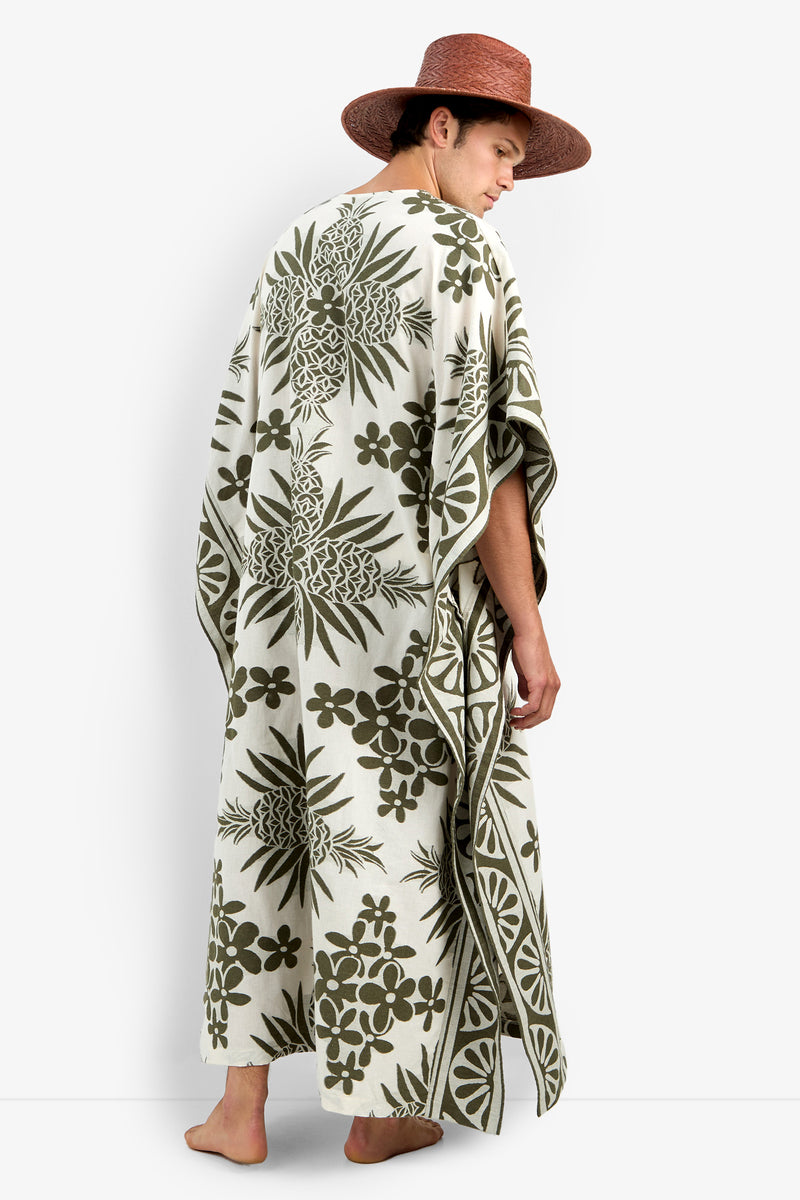 A person stands barefoot, facing away from the camera, wearing the Wailea Caftan—a long, cream-colored garment with green tropical floral and pineapple patterns and a wide-brimmed hat.