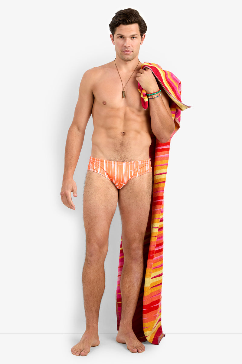 A man stands barefoot against a white background, wearing the Bondi Swim Brief in orange and white stripes. He holds a colorful striped beach towel over his shoulder and accessorizes with bracelets and a necklace.