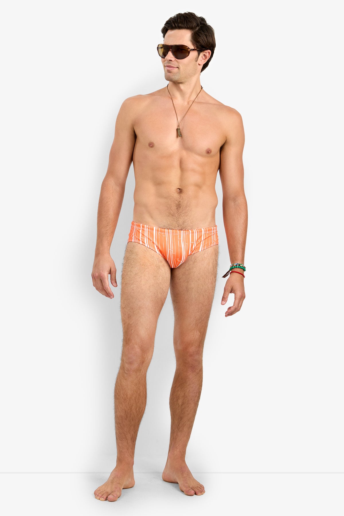 A man stands barefoot against a plain white background, looking slightly to the side while wearing sunglasses, jewelry, and the Bondi Swim Brief in orange and white stripes.
