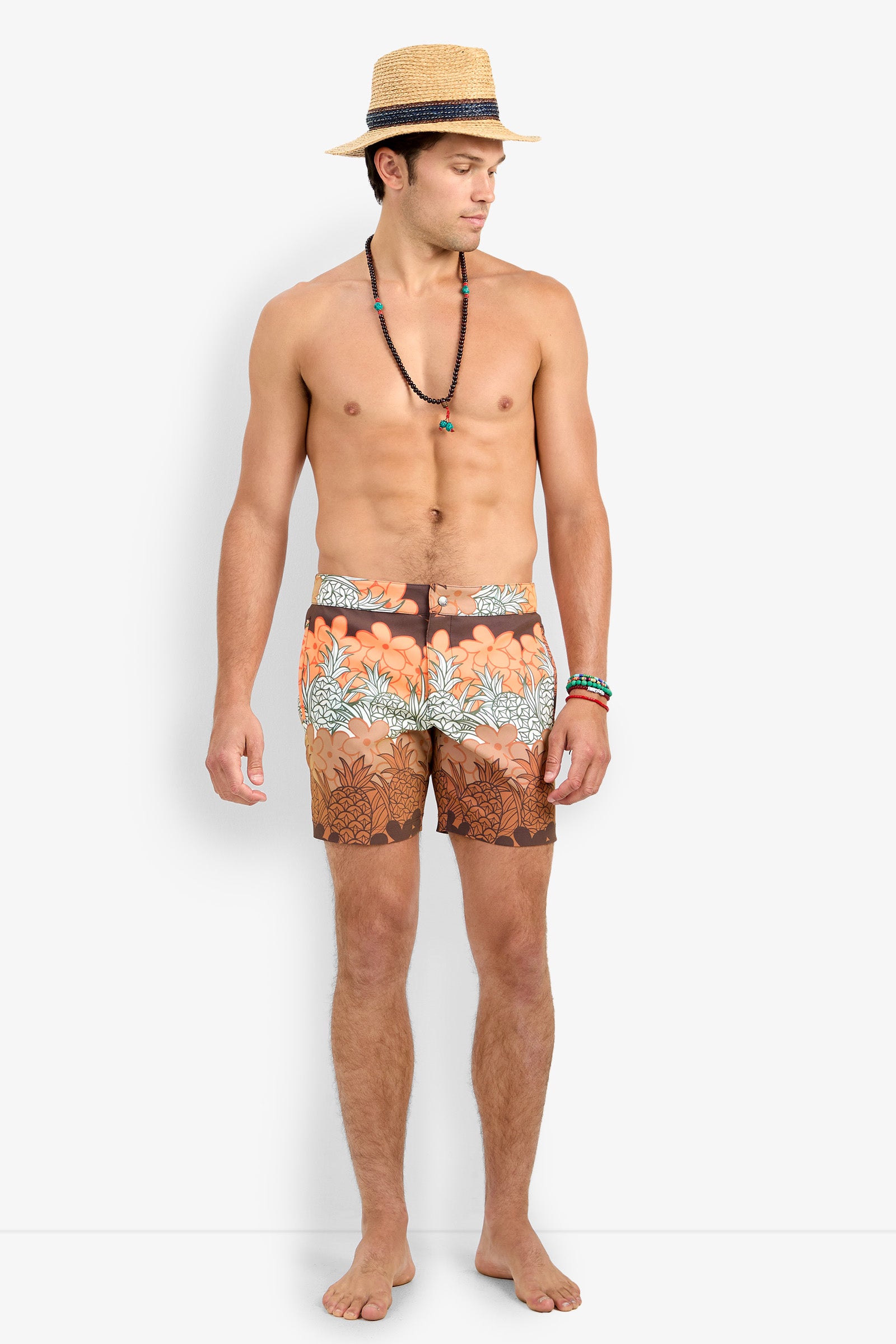 A man stands barefoot, wearing the Honolulu Board Short—orange with brown and white floral patterns—a beaded necklace, and a straw hat. Shirtless, he looks left against a plain white background.