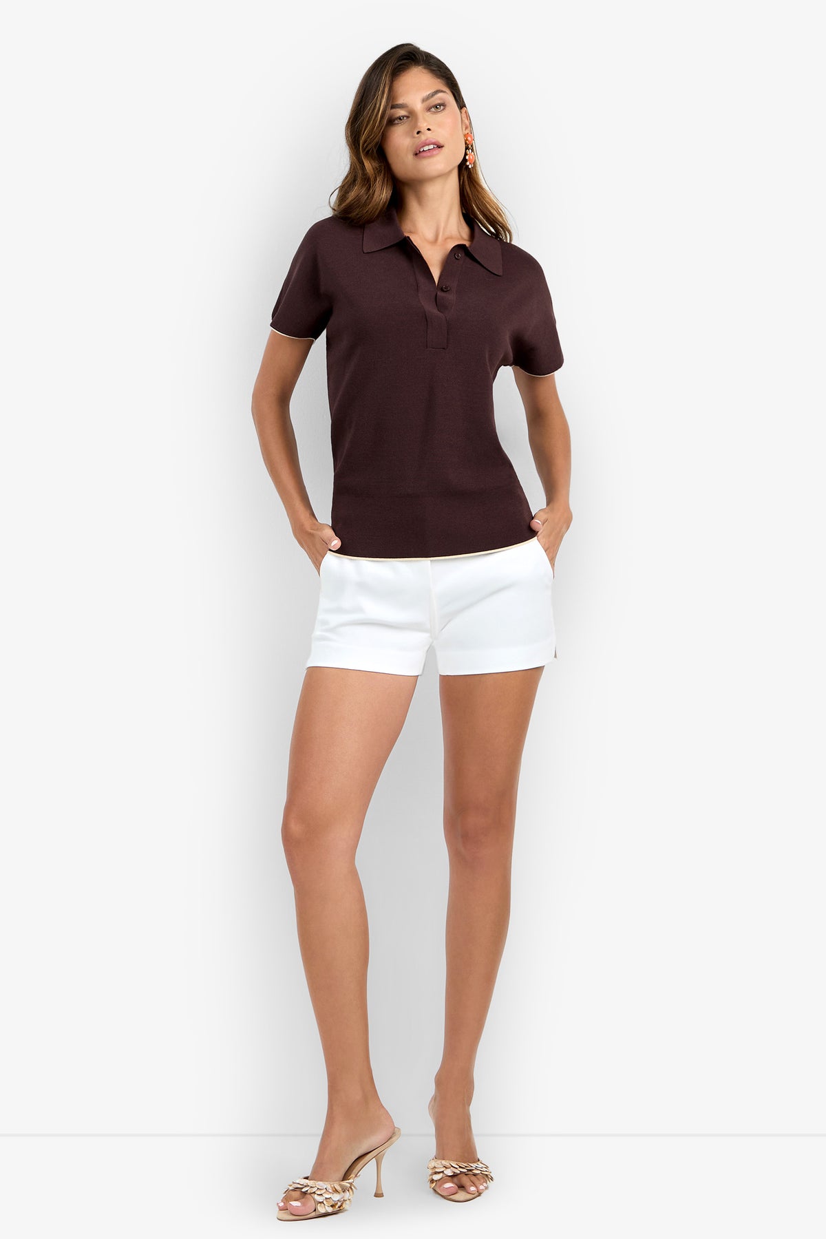 A woman stands with her hands in her pockets, wearing the Hilo Pullover—a short-sleeved dark brown collared shirt—paired with white shorts and crisscross-strap heels, set against a plain light background.