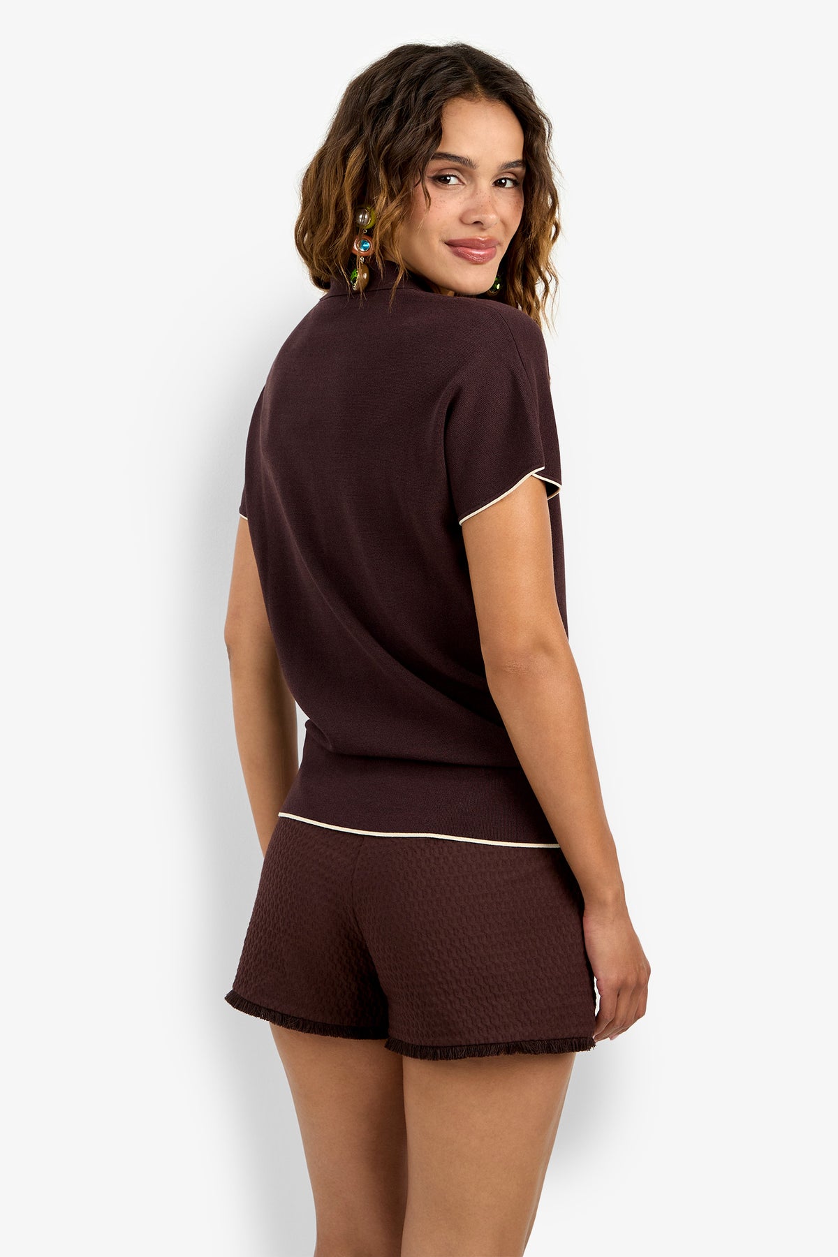 A woman with wavy brown hair, seen from behind and looking over her shoulder, wears the Hilo Pullover—a short-sleeved, dark brown top—paired with matching textured shorts with light trim against a plain white background.