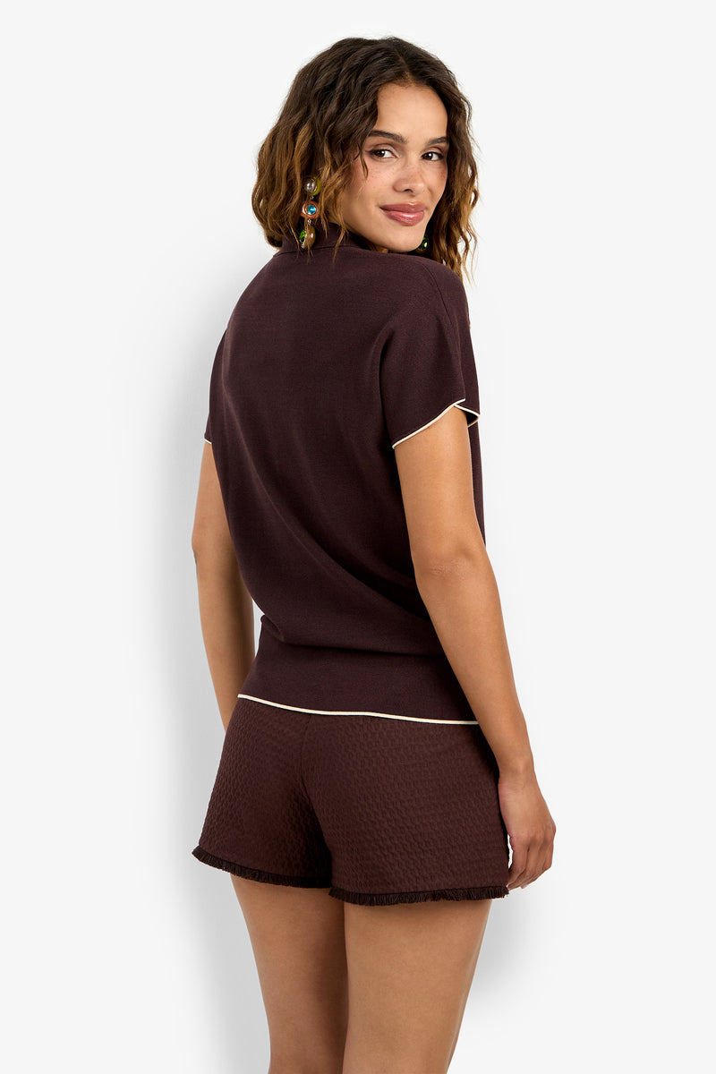 A woman with wavy brown hair, seen from behind and looking over her shoulder, wears the Hilo Pullover—a short-sleeved, dark brown top—paired with matching textured shorts with light trim against a plain white background.