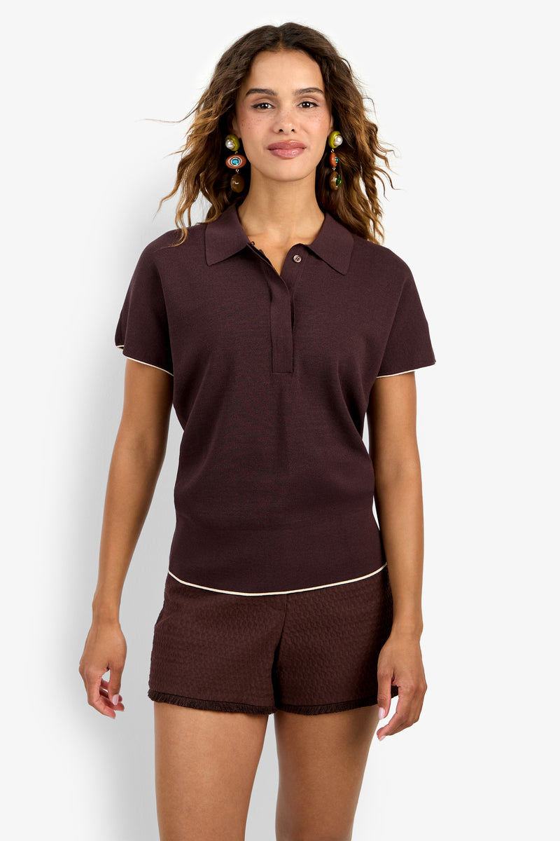 Wearing the Hilo Pullover, a person with wavy hair stands against a plain white background, accessorized with colorful dangling earrings and brown textured shorts.