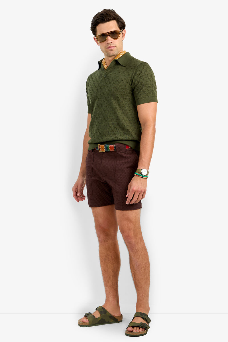 A man in sunglasses, the Waylen Short Sleeve Polo in olive green, patterned brown shorts, a colorful belt, watch, and green camo sandals stands against a plain white background.