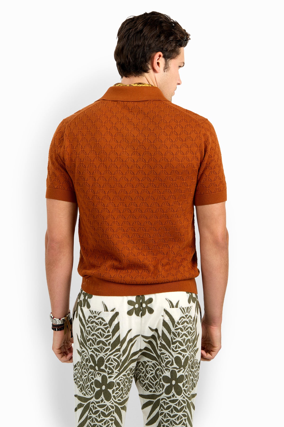 A man stands facing away, wearing the Waylen Short Sleeve Polo in rust and high-waisted white pants with bold green floral and geometric patterns.