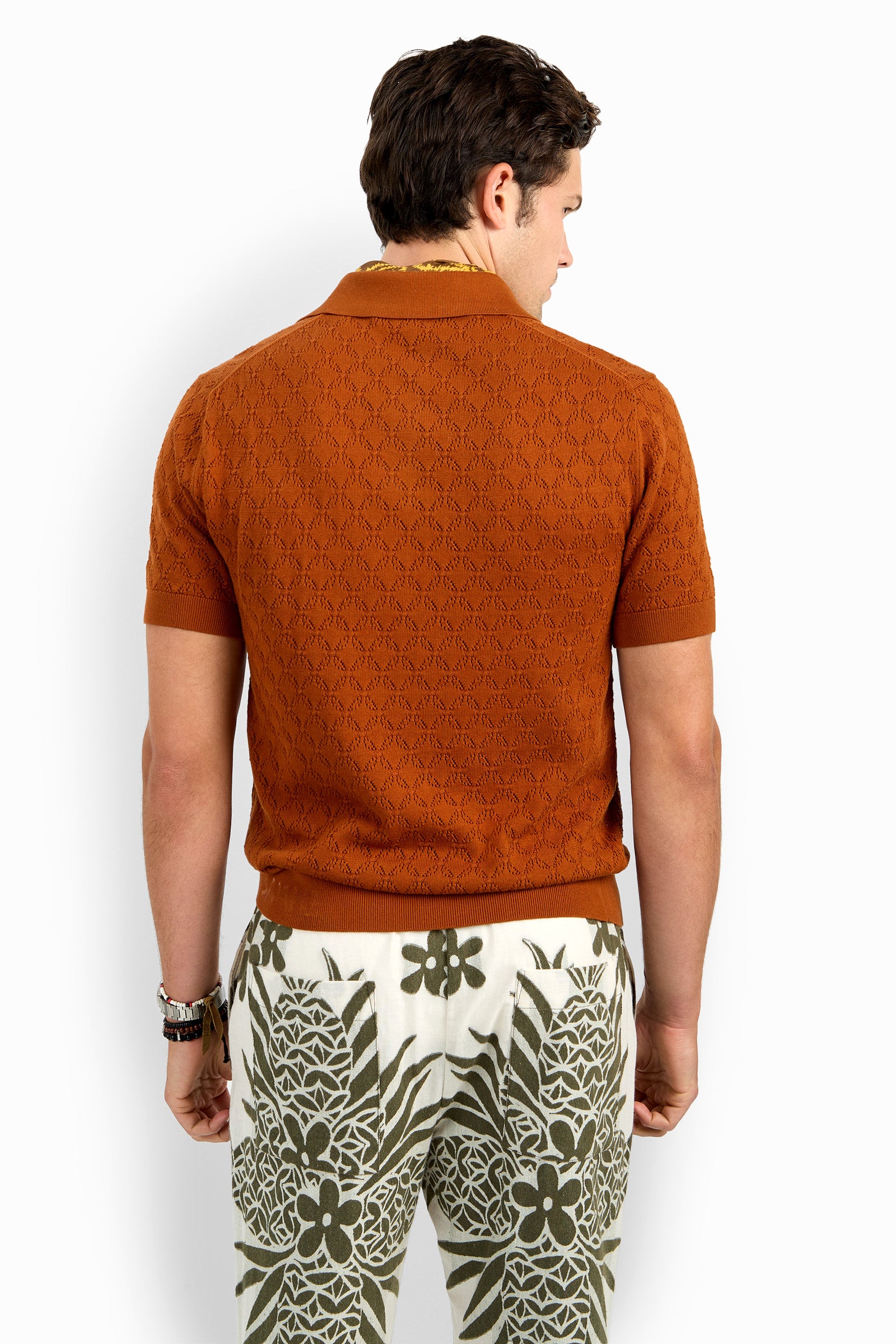 A man stands facing away, wearing the Waylen Short Sleeve Polo in rust and high-waisted white pants with bold green floral and geometric patterns.