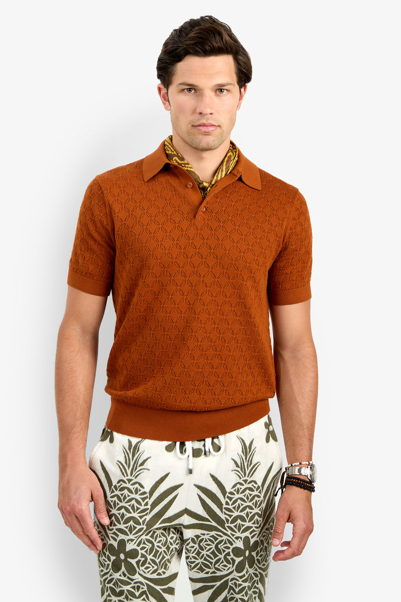A man with short brown hair and a neutral expression stands against a plain background wearing the Waylen Short Sleeve Polo in orange, a gold patterned scarf, and cream pants featuring green pineapple and floral prints.