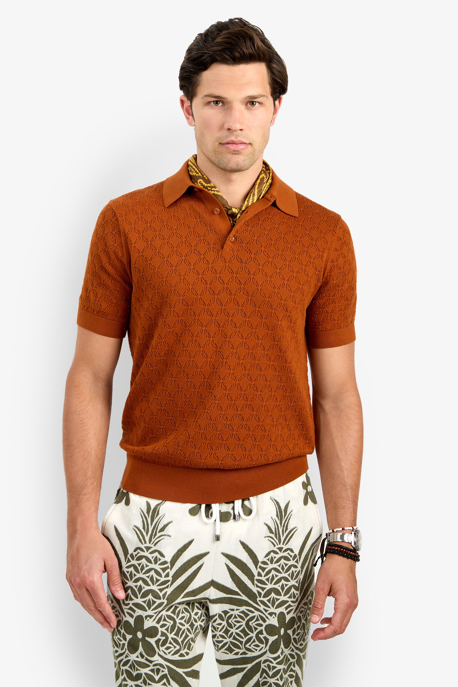 A man with short brown hair and a neutral expression stands against a plain background wearing the Waylen Short Sleeve Polo in orange, a gold patterned scarf, and cream pants featuring green pineapple and floral prints.