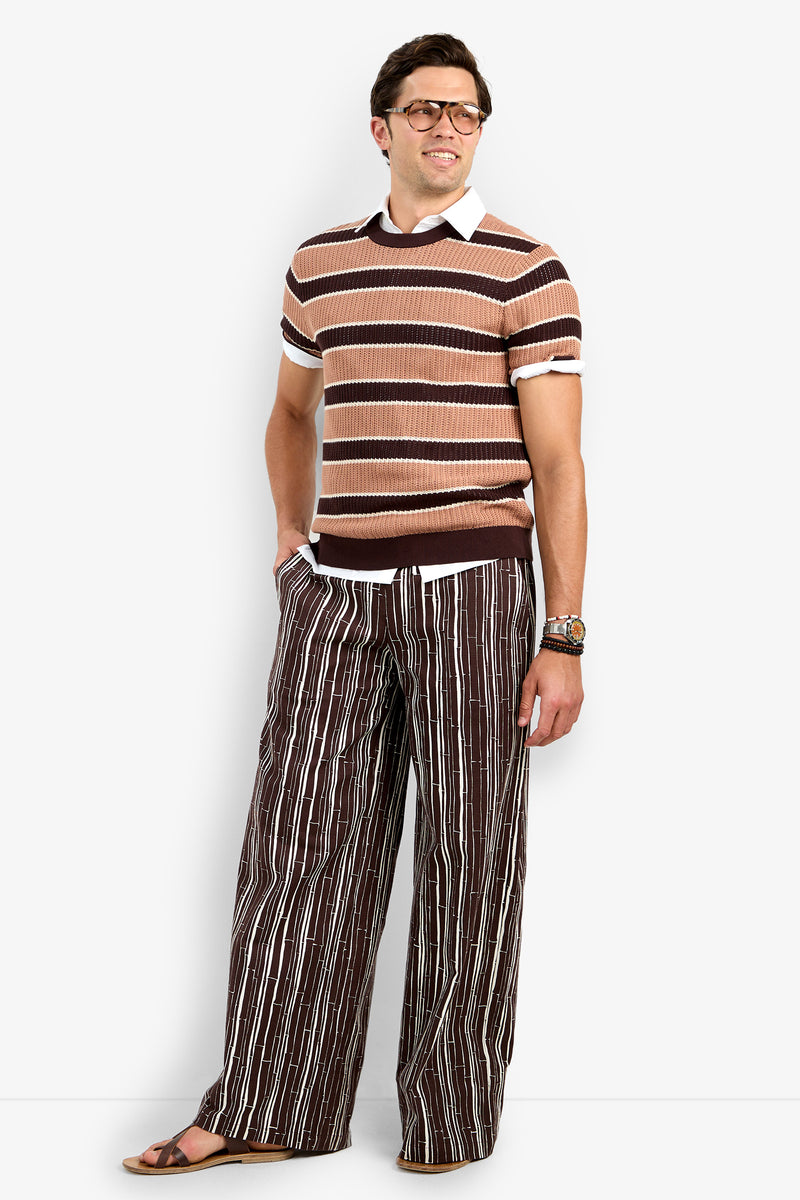 A man wearing glasses and the Carlos Short Sleeve Crewneck over a collared shirt, with wide-leg striped pants, sandals, and a bracelet, stands smiling with one hand in his pocket against a plain white background.