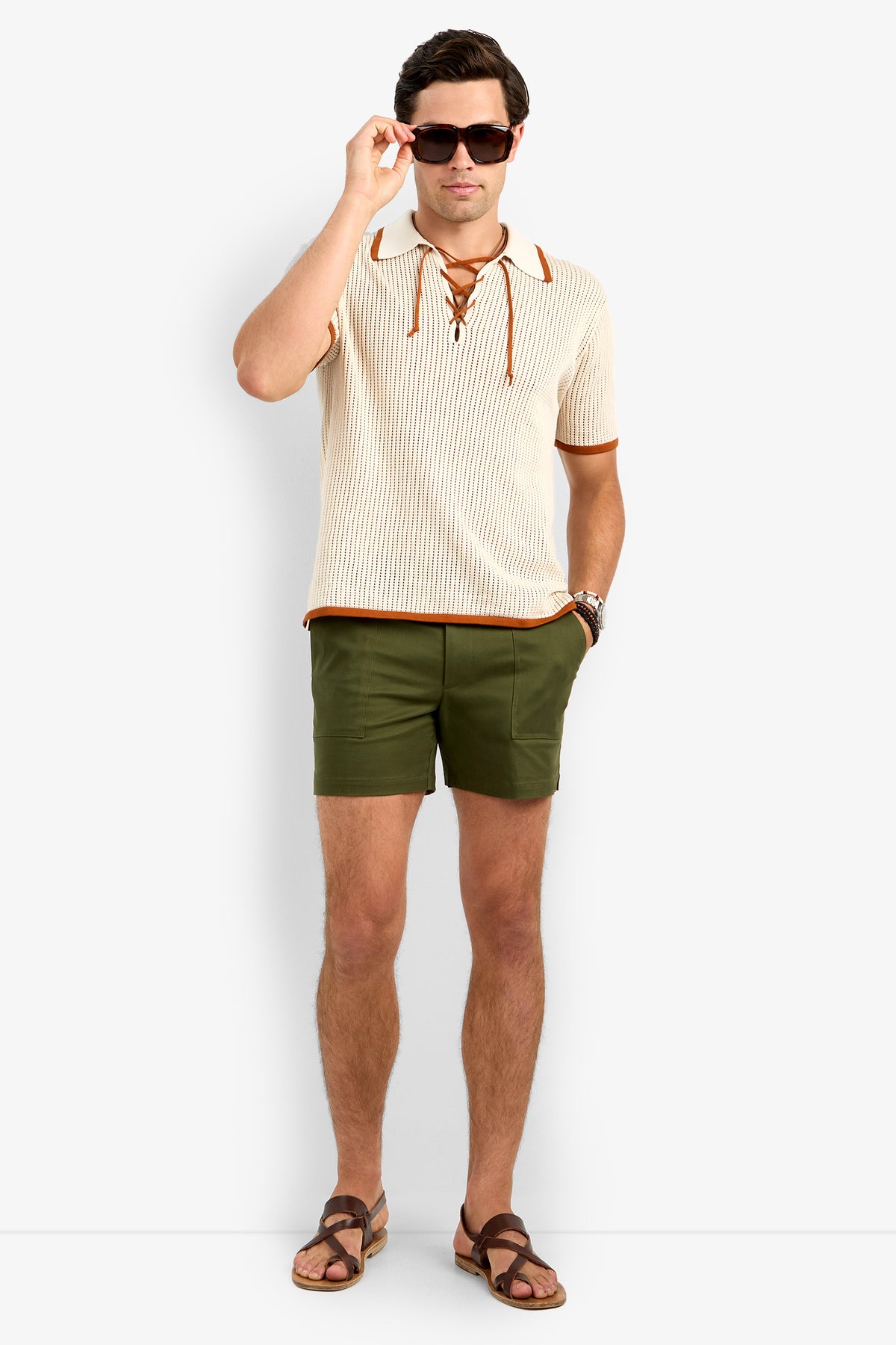A man in sunglasses, the cream Simon Lace Up Polo with brown trim, olive green shorts, and brown sandals stands against a plain white background.