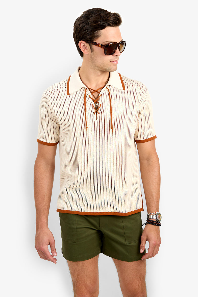 A man stands against a plain light background, looking slightly to the side, wearing dark sunglasses, olive green shorts, and the Simon Lace Up Polo in cream with short sleeves and lace-up detail.