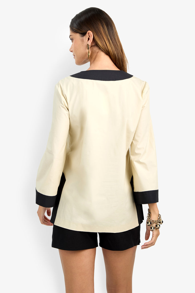 A woman with long brown hair, gold earrings, and black shorts models the DAMETRIA TOP—cream with black collar and cuffs—standing against a plain white background, facing away from the camera.