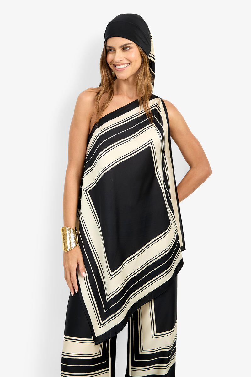 A woman smiles while posing against a plain white background, wearing the DIANN TOP—a black and beige geometric-patterned one-shoulder piece—paired with matching pants, a gold cuff bracelet, and a black headscarf.