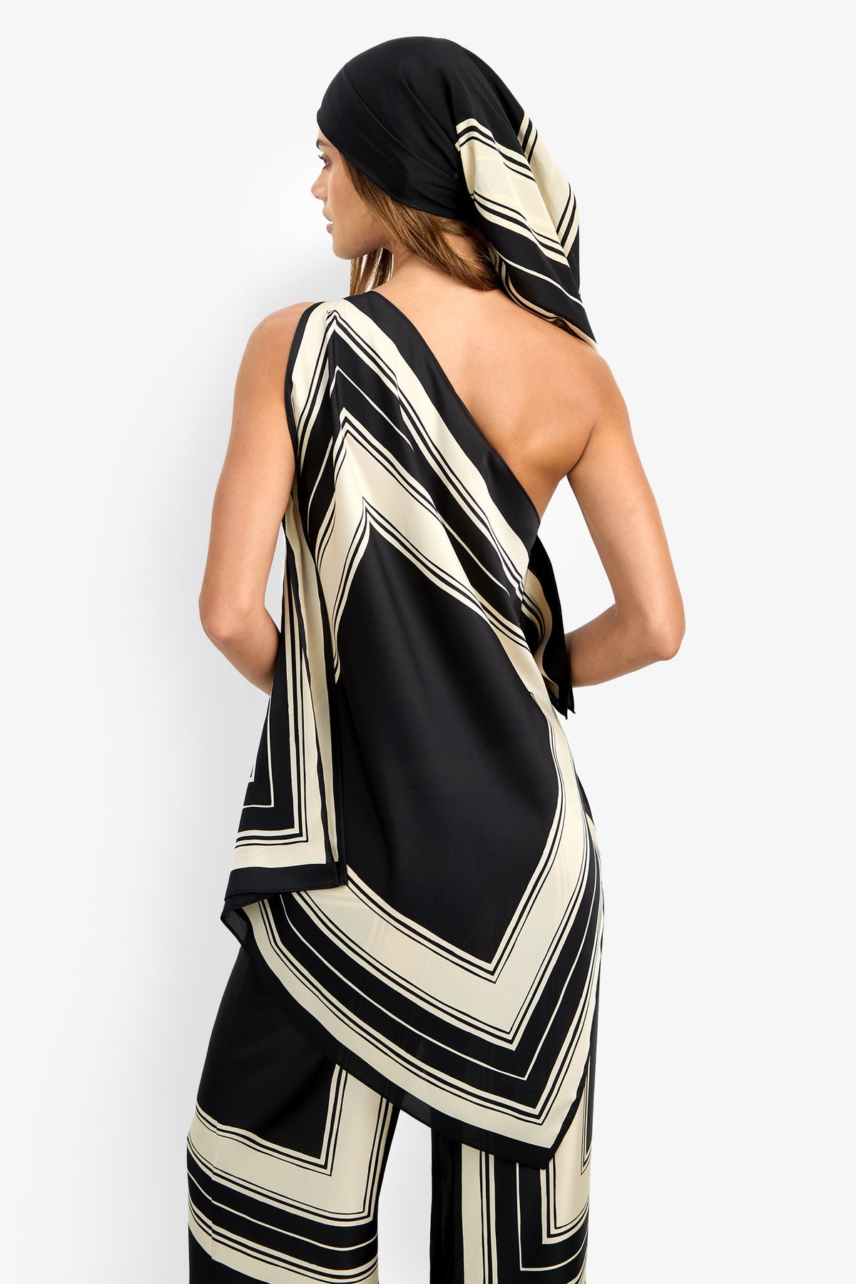 A woman faces away, wearing the DIANN TOP—a black and cream geometric-patterned, one-shoulder piece—paired with matching pants and a headscarf. Her right shoulder is bare, and her long hair flows beneath the scarf.