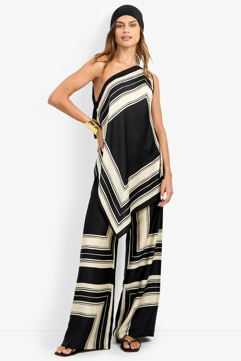 A woman stands against a white background wearing the DIANN TOP, a black and cream geometric one-shoulder asymmetrical top with matching wide-leg pants, black sandals, a gold cuff bracelet, and a black headscarf.