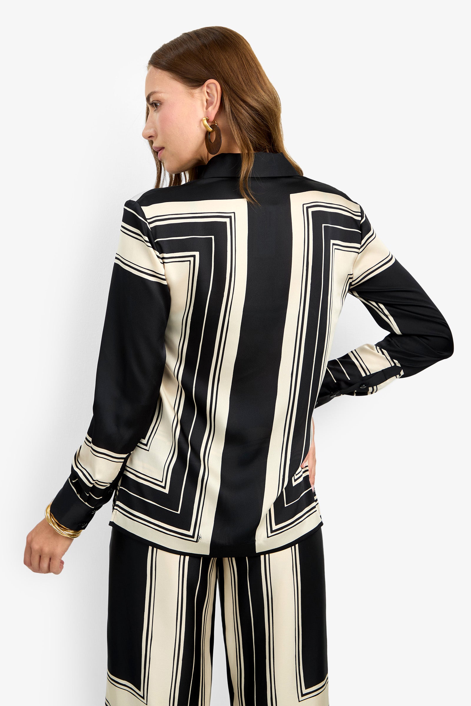 Woman with light brown hair stands with her back to the camera, looking over her shoulder while wearing the BRISTLECONE TOP, a black and white geometric-patterned long-sleeve blouse paired with matching pants.