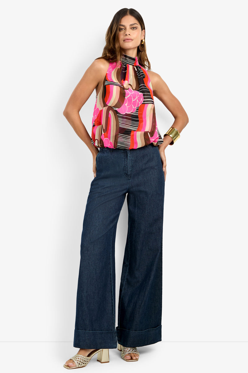 A woman stands confidently wearing the wide-leg DILLAN PANT in dark denim, paired with a colorful sleeveless halter top, heeled sandals, hoop earrings, and a large gold cuff bracelet.