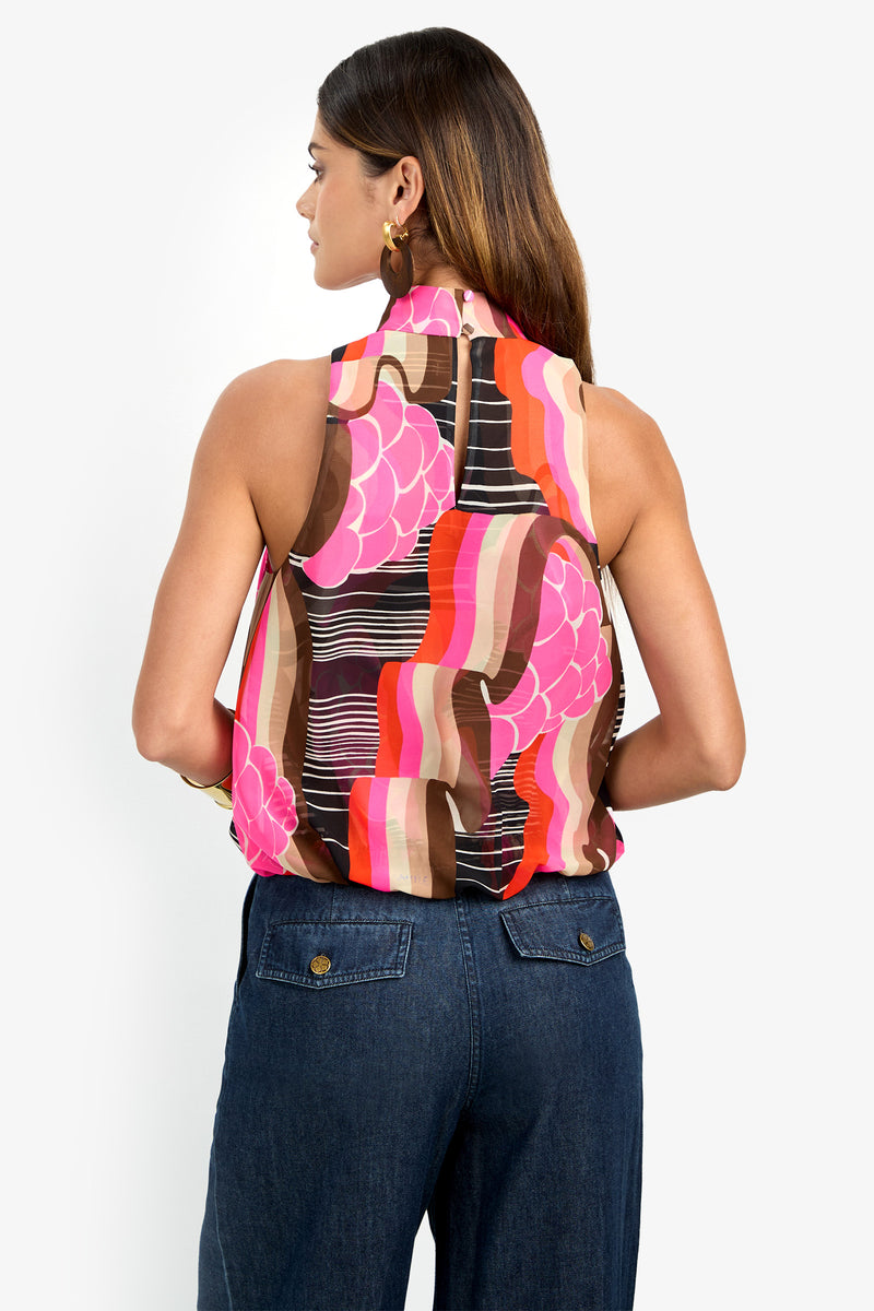 A woman with straight brown hair and gold hoop earrings faces away, wearing the sleeveless CORTONA TOP in a bold pink, red, brown, and white abstract print, paired with high-waisted blue jeans.