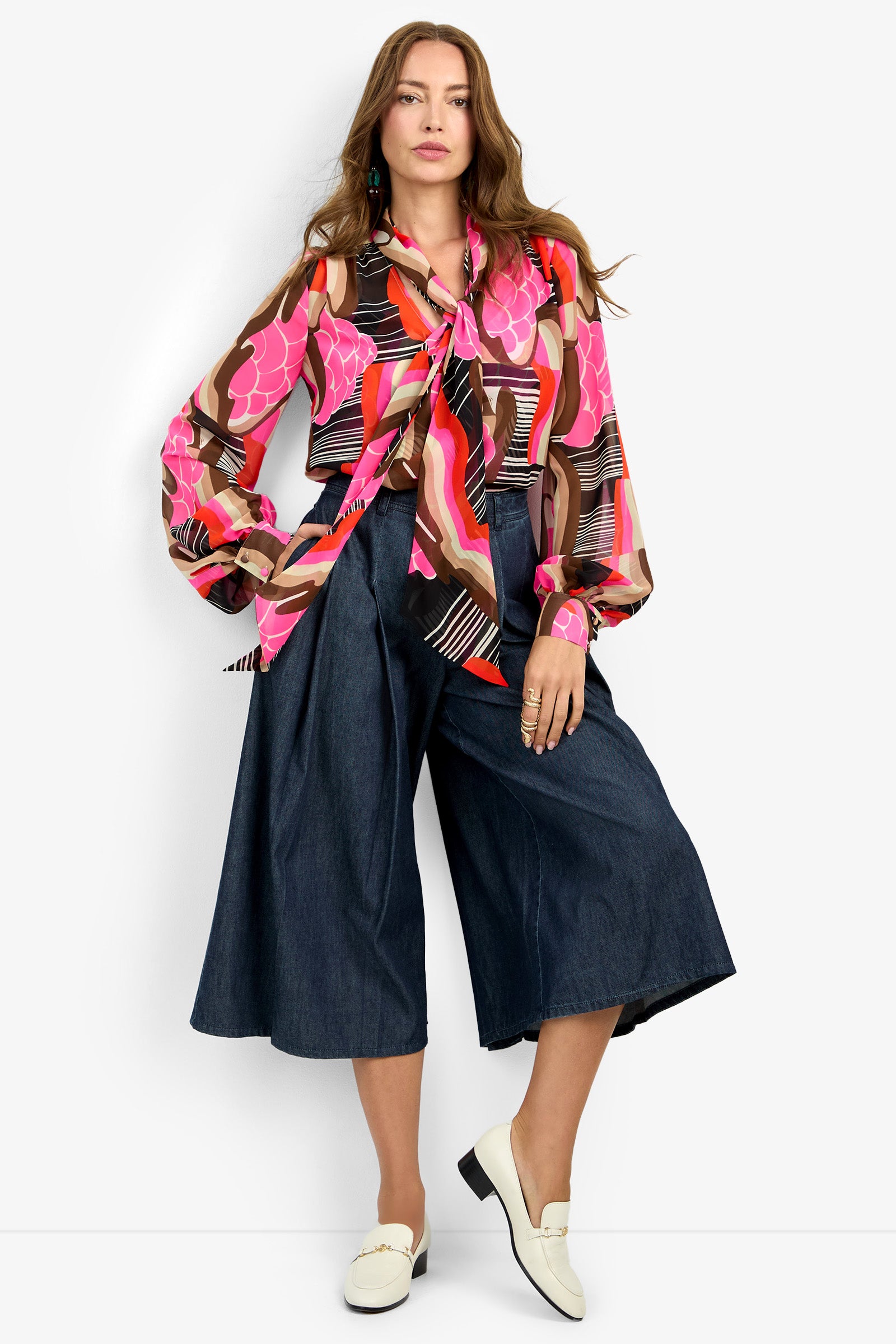 A woman poses confidently against a white background wearing the PIENZA TOP, a colorful, patterned blouse with a large bow, paired with wide-leg dark denim culottes and white loafers.