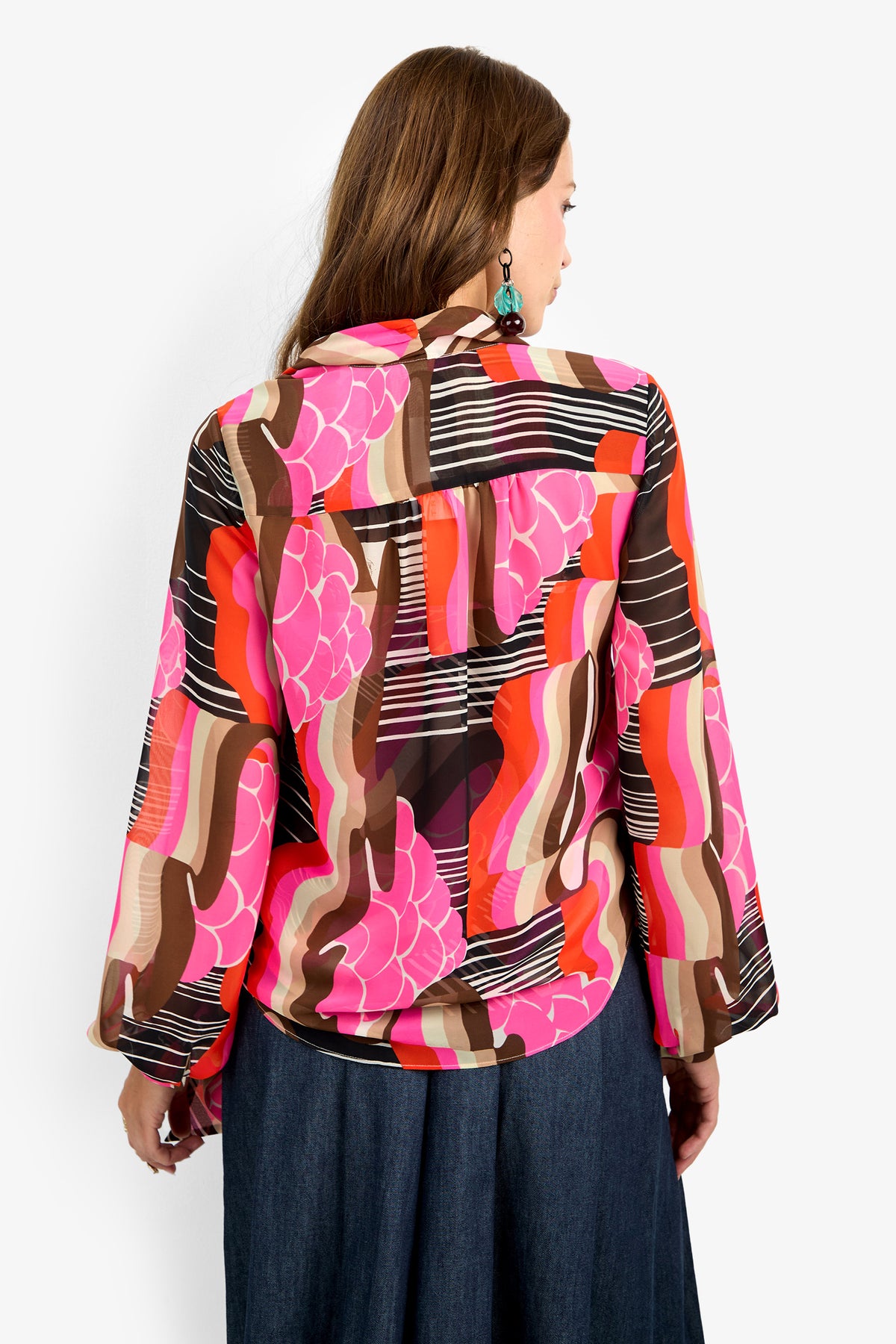 A woman with long brown hair, seen from behind, wears the PIENZA TOP—a colorful abstract blouse in pink, orange, brown, and black—paired with a denim skirt and statement earrings against a plain white background.