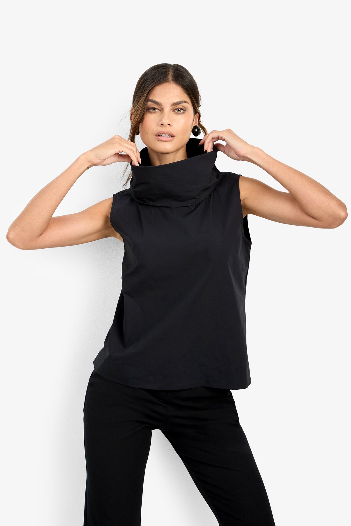 A woman poses against a white backdrop, wearing the CASTIGLIONE TOP—a sleeveless black design with a wide, structured cowl neck—paired with black pants. She adjusts the collar with both hands and looks confidently at the camera.