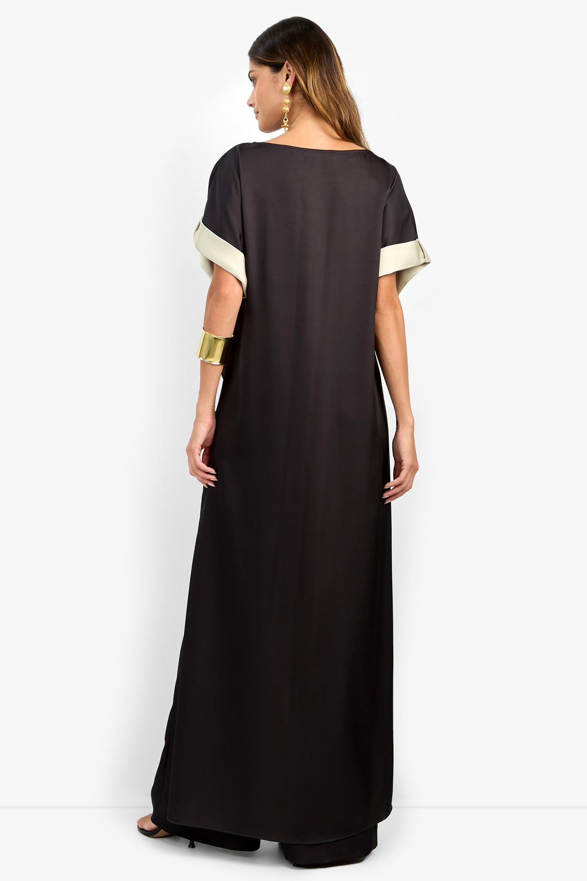 A woman with long brown hair wears the MATTIAH TOP, a black short-sleeve top with light trim on the cuffs. She accessorizes with gold earrings and a wide gold cuff bracelet, shown from the back.