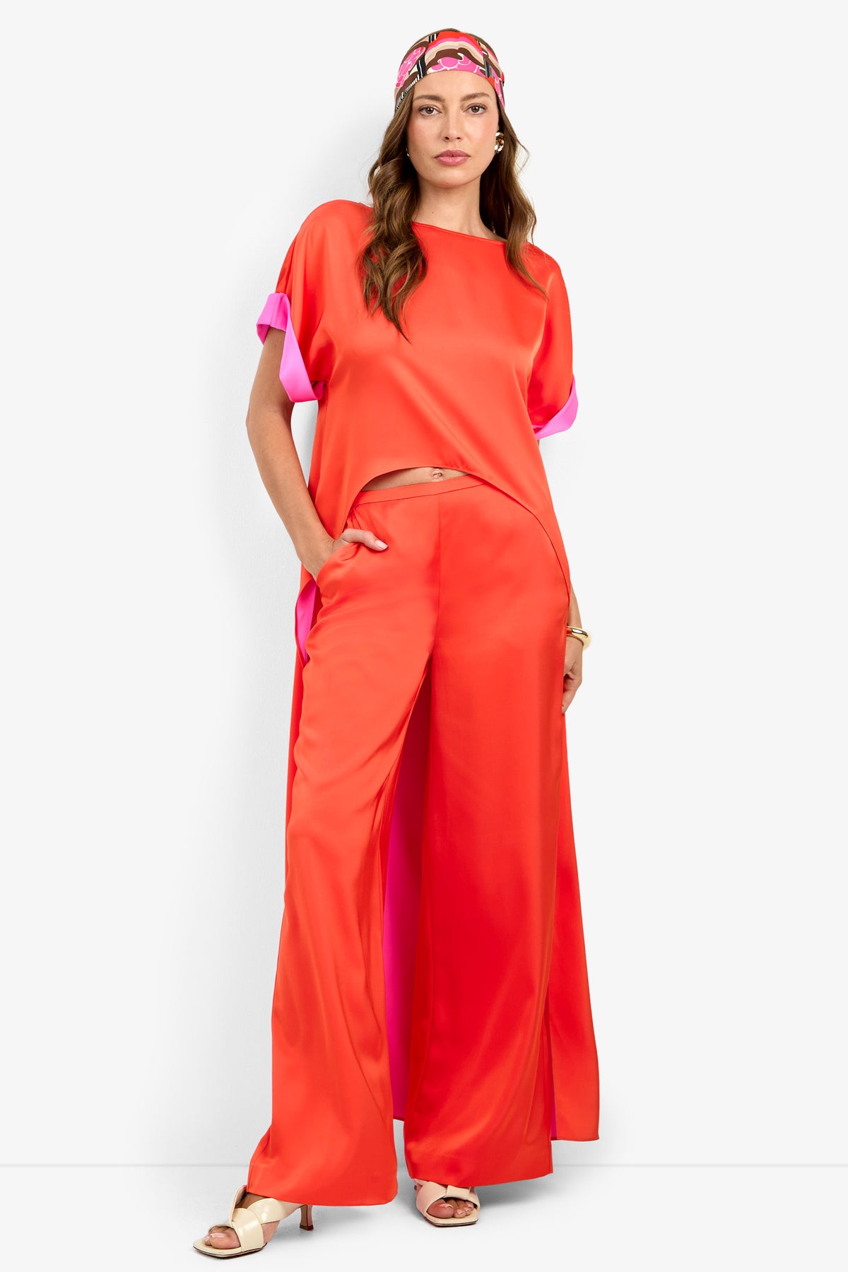 A woman poses with one hand in her pocket, wearing the loose-fit MATTIAH TOP in bright red satin with pink details and matching wide-leg pants, styled with a colorful headband and white heeled sandals.