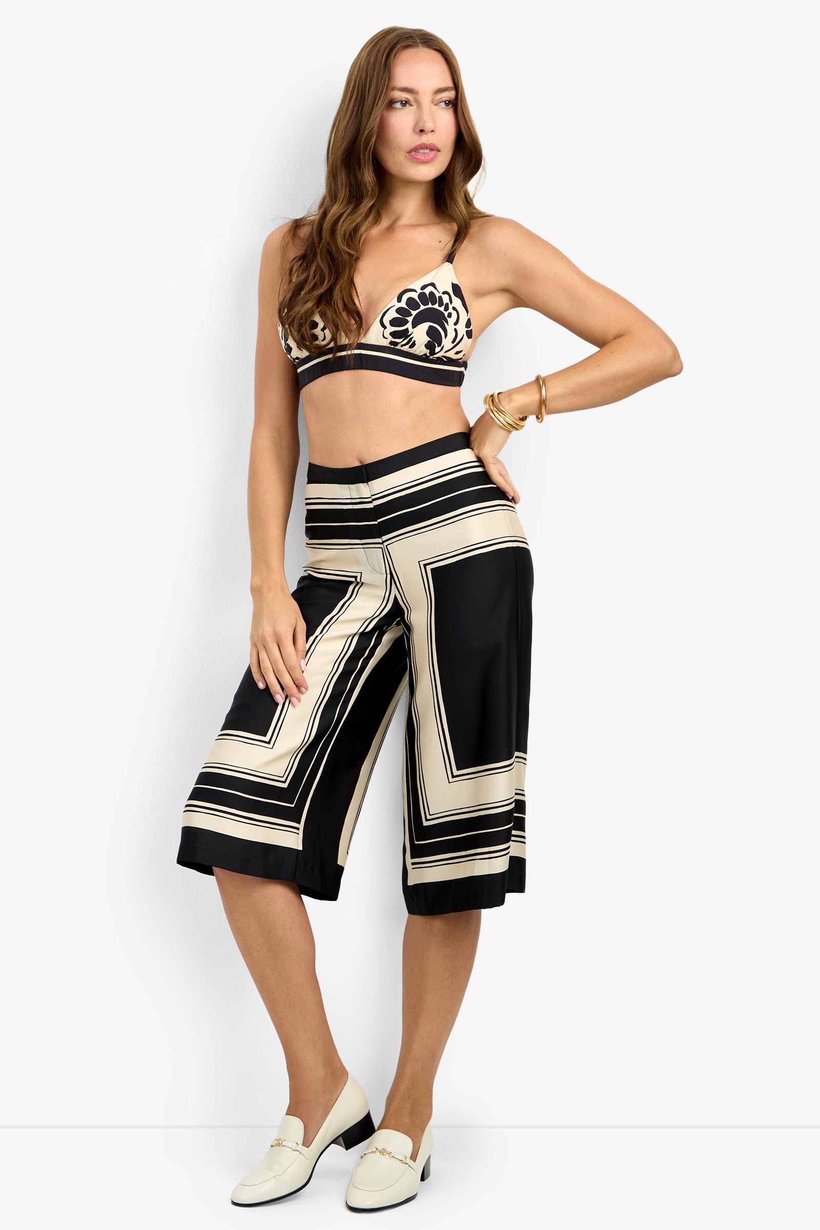 A woman wearing the BLANCA TOP, wide-leg culotte pants with black and cream geometric designs, cream loafers, and gold bracelets poses with one hand on her hip.