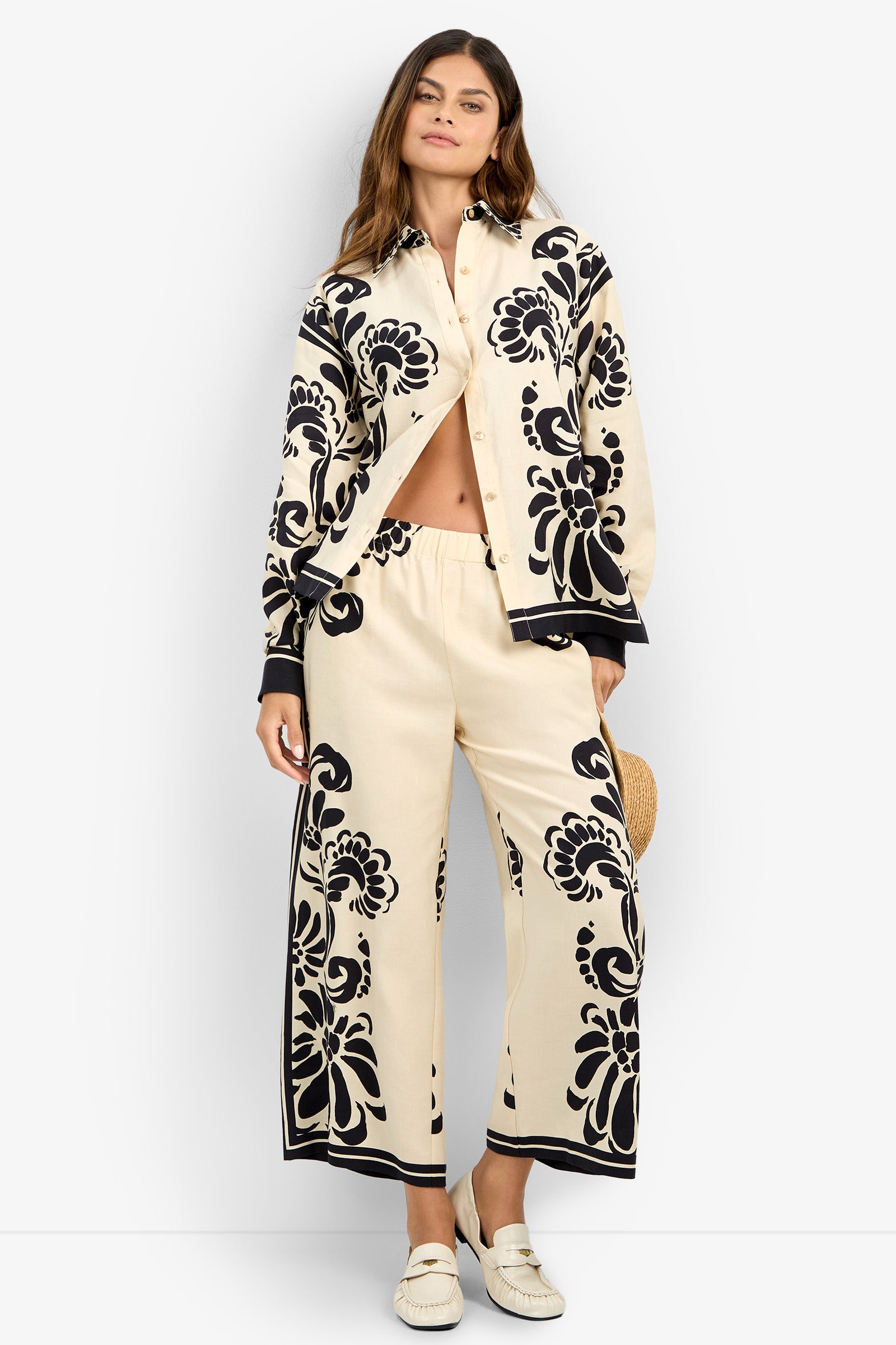 A woman wears the NIVARA TOP, a cream shirt with bold black floral patterns, paired with matching wide-leg pants. She stands against a white background, holding a straw hat and wearing white loafers.