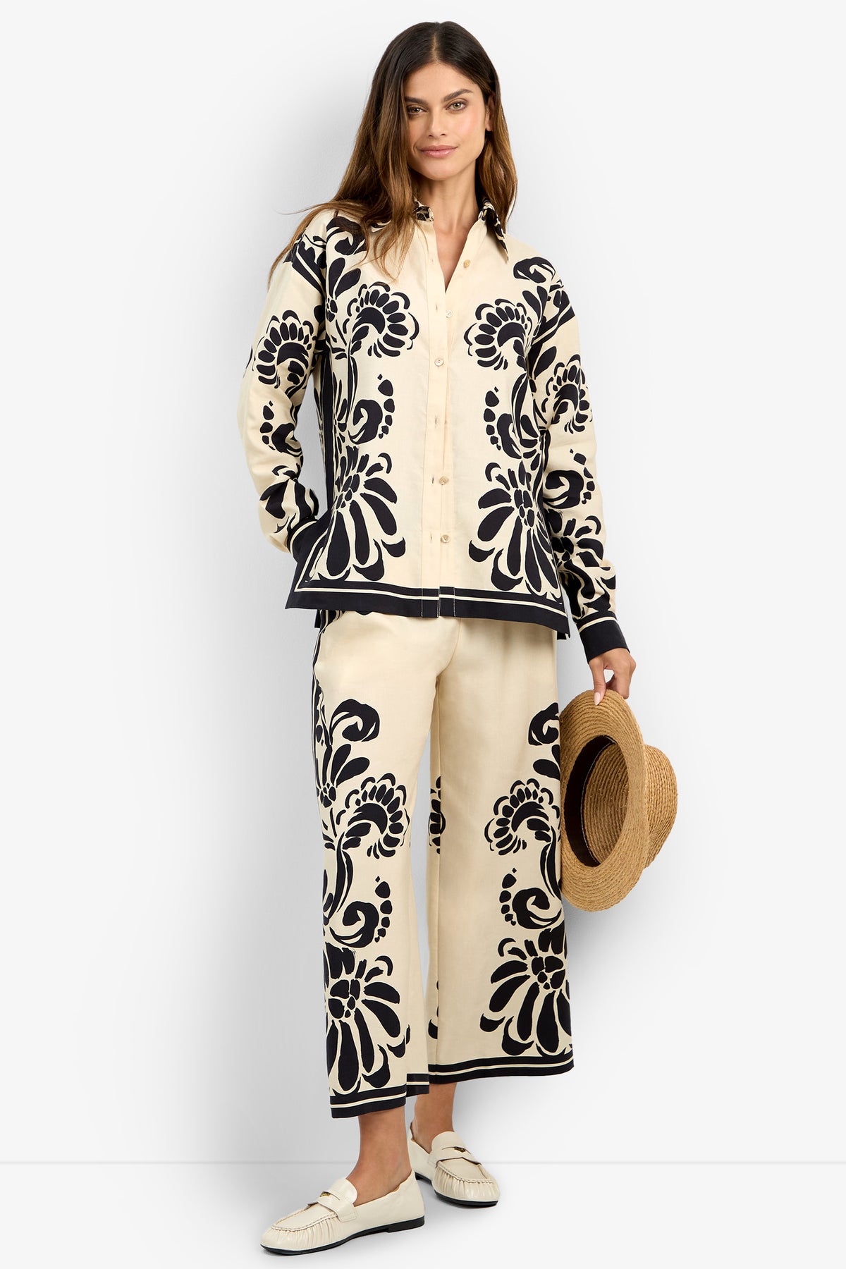 A woman with long hair and a neutral expression wears the NIVARA TOP—a cream-colored shirt with bold black floral patterns—paired with matching wide-leg pants, white loafers, and holds a straw hat by her side.