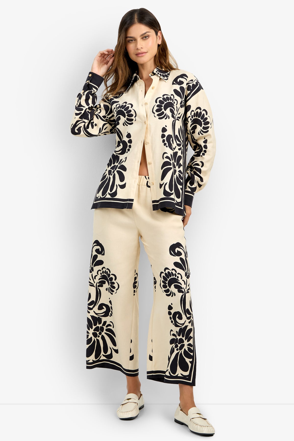 A woman wearing the NIVARA TOP, a cream matching set with bold black floral patterns and wide-leg pants, paired with white loafers, stands against a plain white background.