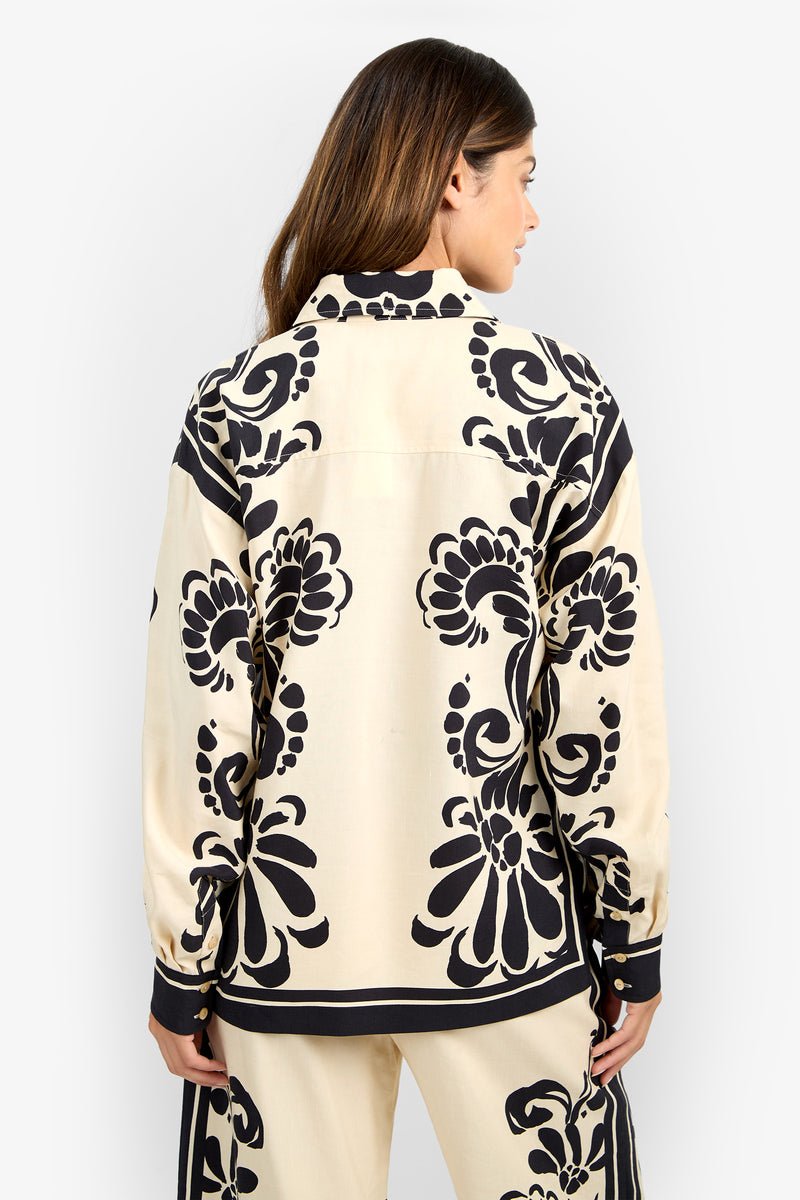 A woman with long brown hair is seen from the back wearing the NIVARA TOP, a cream-colored shirt featuring bold black floral patterns, paired with matching pants against a plain white background.