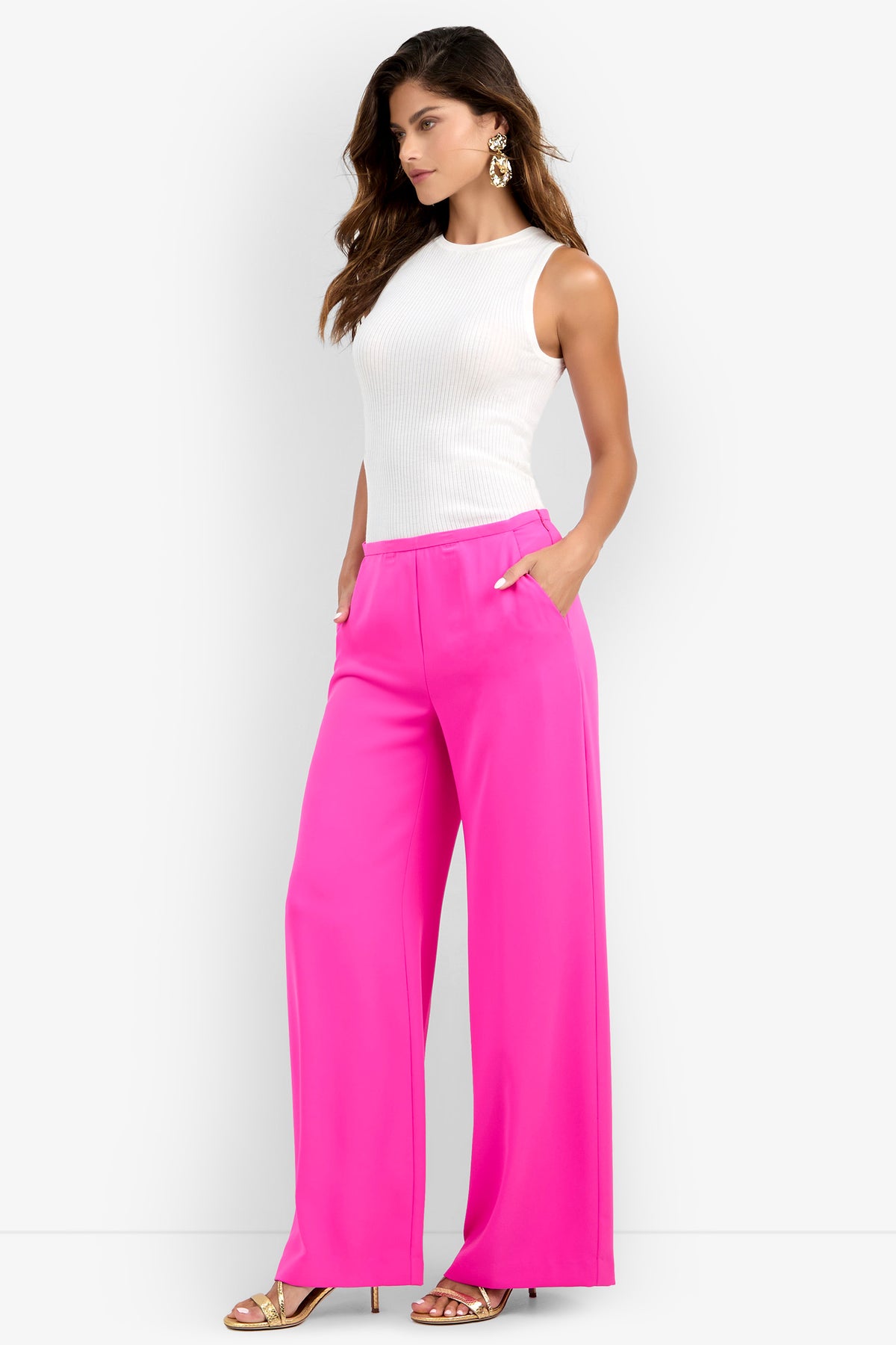 A woman wearing the Lyndi Top, a sleeveless white top, paired with bright pink wide-leg pants, gold earrings, and gold heeled sandals, stands with her hands in her pockets against a plain white background.