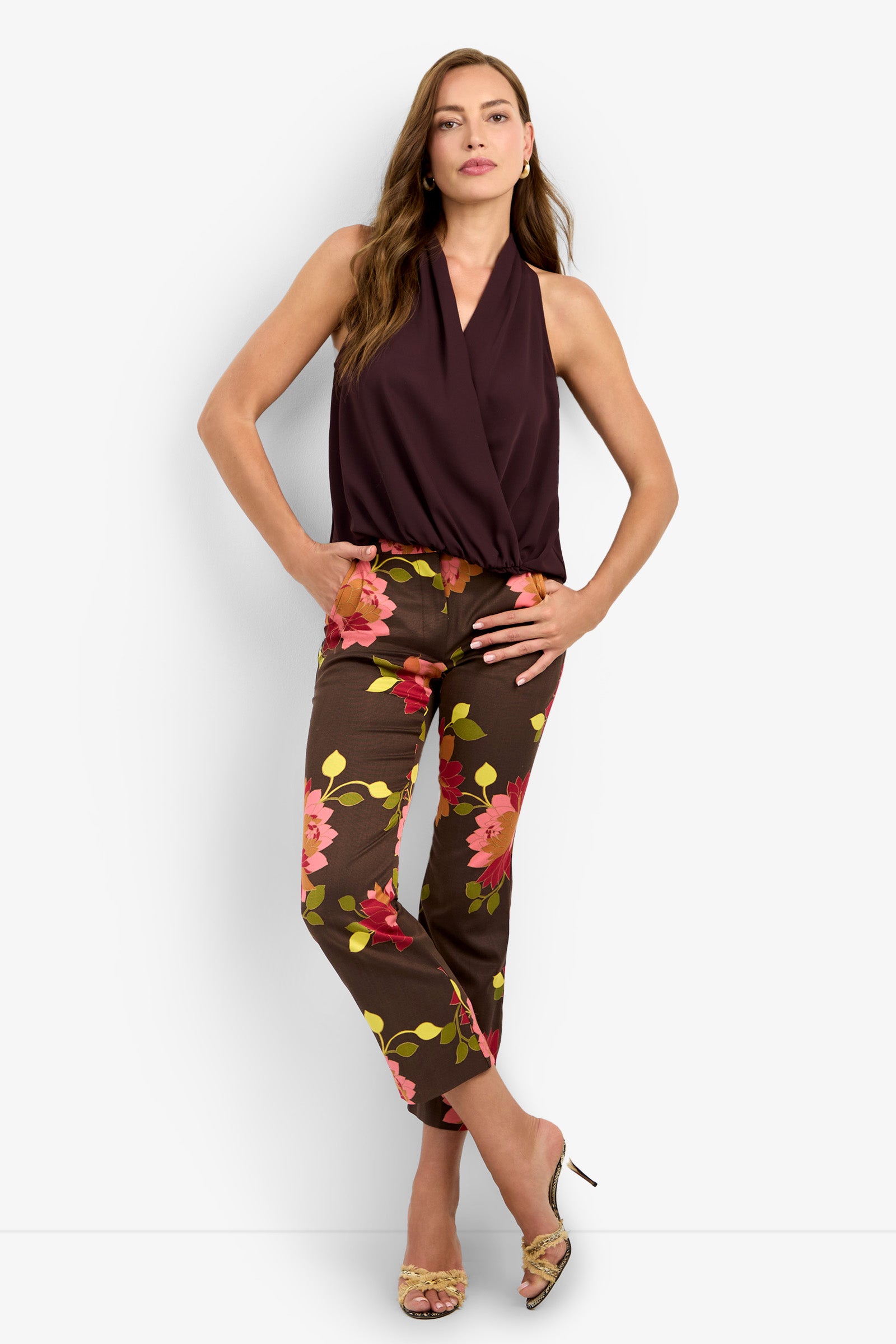 A woman stands confidently wearing the LUCCA TOP, a sleeveless deep burgundy wrap top, paired with brown ankle-length floral pants. She has long brown hair and open-toe heels.