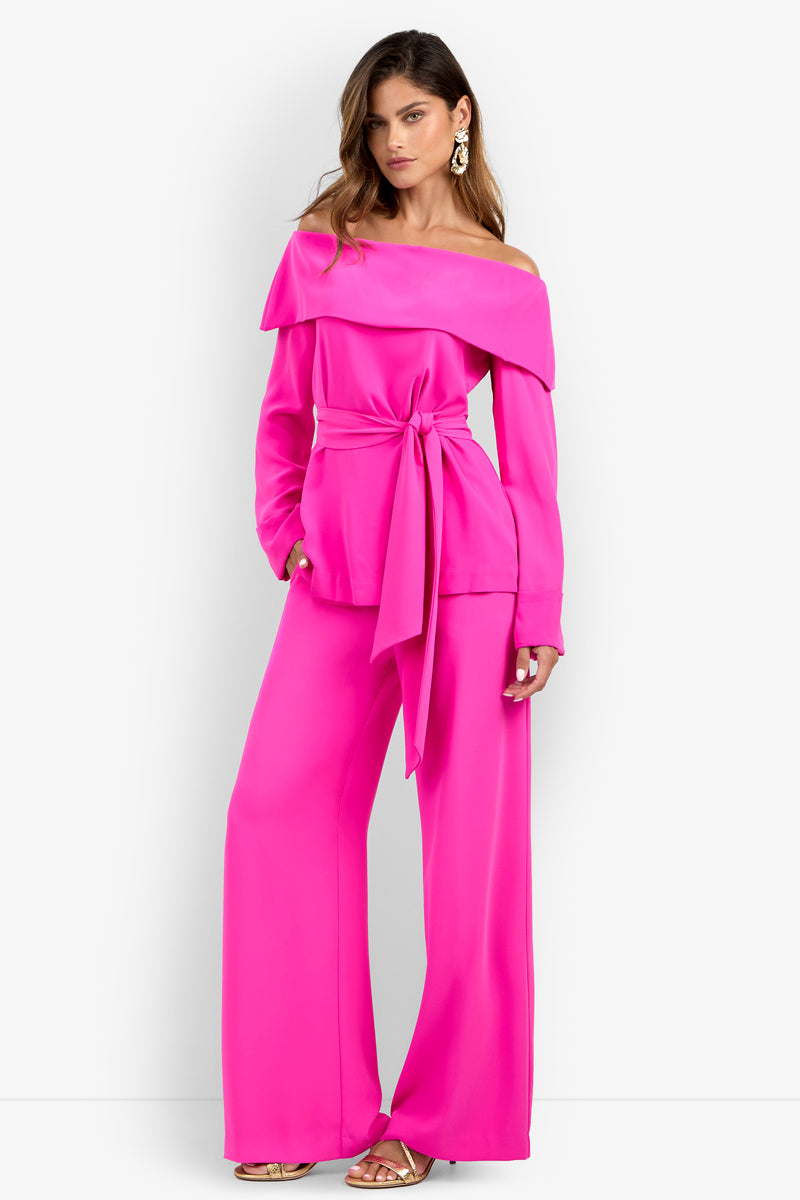 A woman wearing the Lyndi Top in bright pink, styled off-shoulder with a belted waist and wide-leg pants, paired with gold heels and statement earrings, stands against a plain white background.