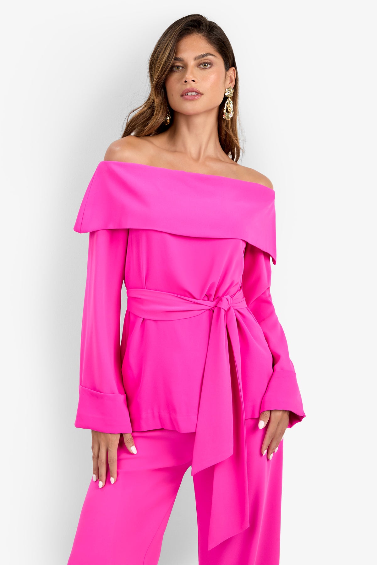 A woman models the Lyndi Top, a bright pink off-the-shoulder style with a wide collar, confidently posing with one hand on her hip against a plain white background.
