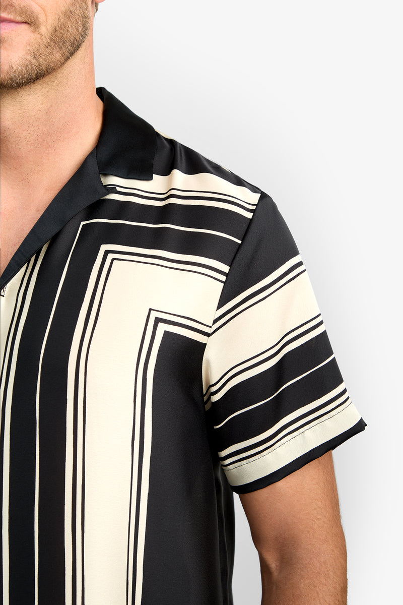A close-up of a person wearing the Felix Camp Shirt, a short-sleeve, black and cream striped collared shirt, photographed from the shoulder up against a plain background.
