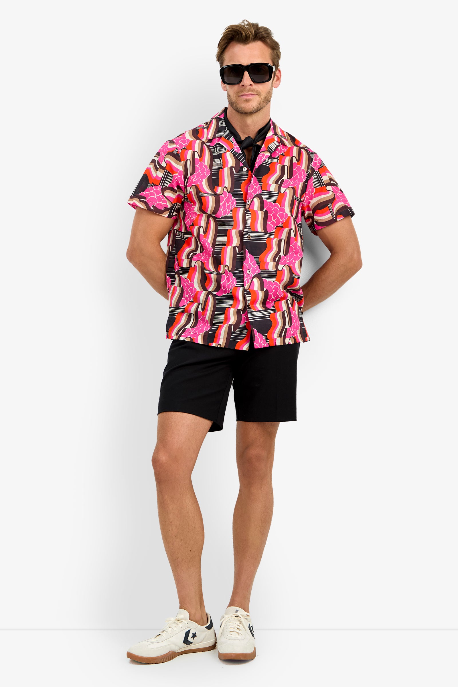 A man wearing black sunglasses, black shorts, white sneakers, and the FELIX CAMP SHIRT with red, pink, and white patterns stands against a plain white background.