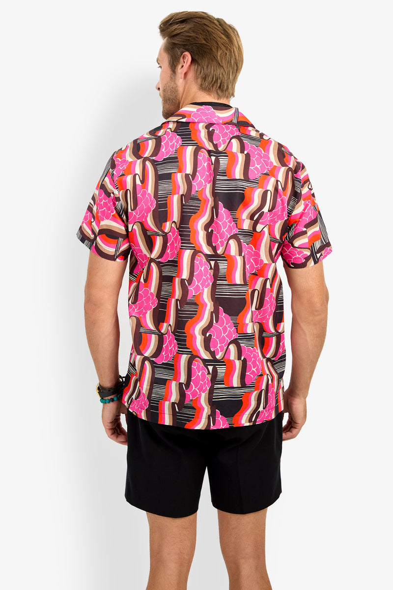 A man faces away, wearing the FELIX CAMP SHIRT—a colorful short-sleeve button-up with a bold abstract pattern in pink, orange, red, black, and white—paired with black shorts.