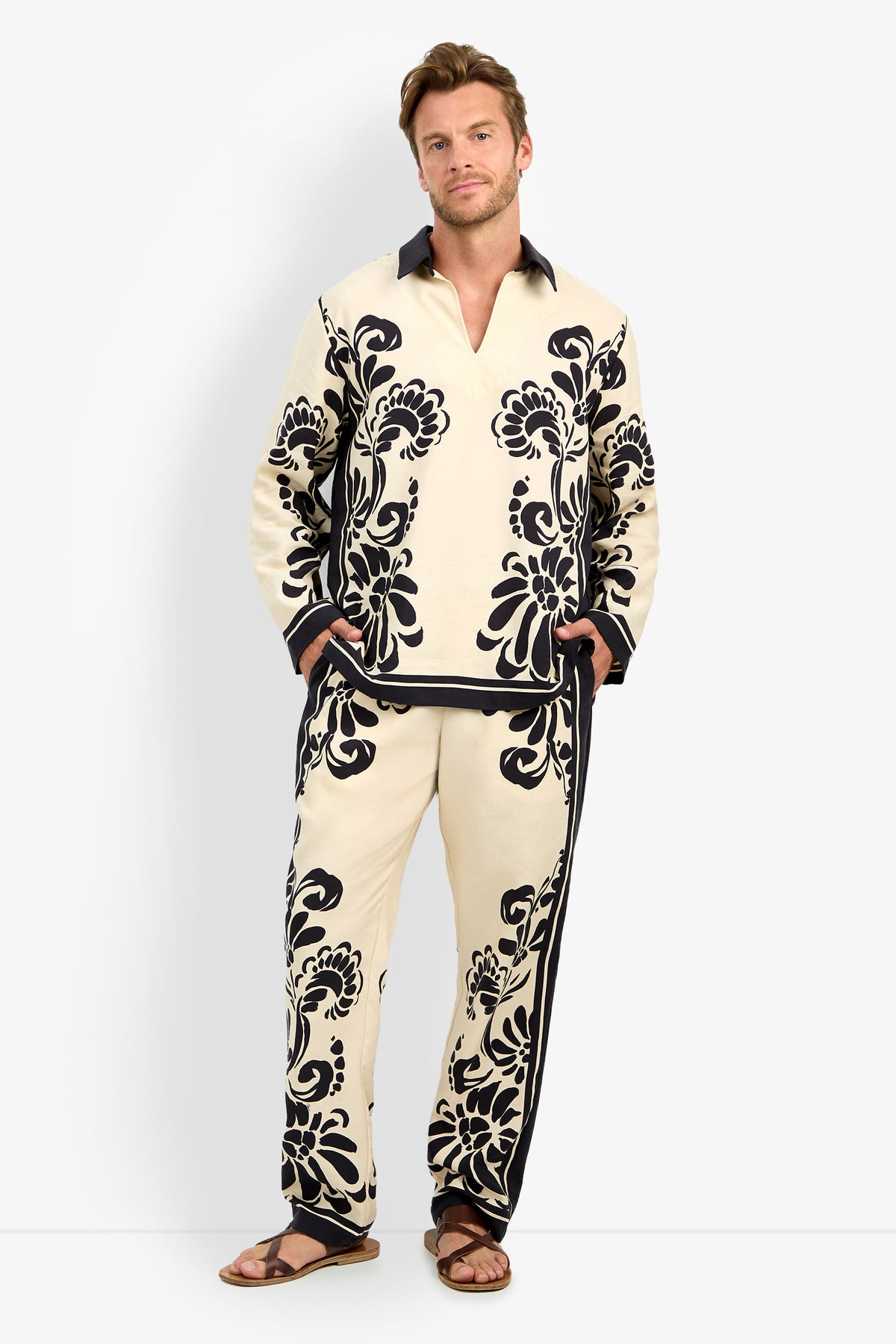 A man stands against a white background wearing the HOLMES PULLOVER in Tivoli, featuring bold black floral patterns and a matching set, paired with brown sandals and his hands in his pockets.
