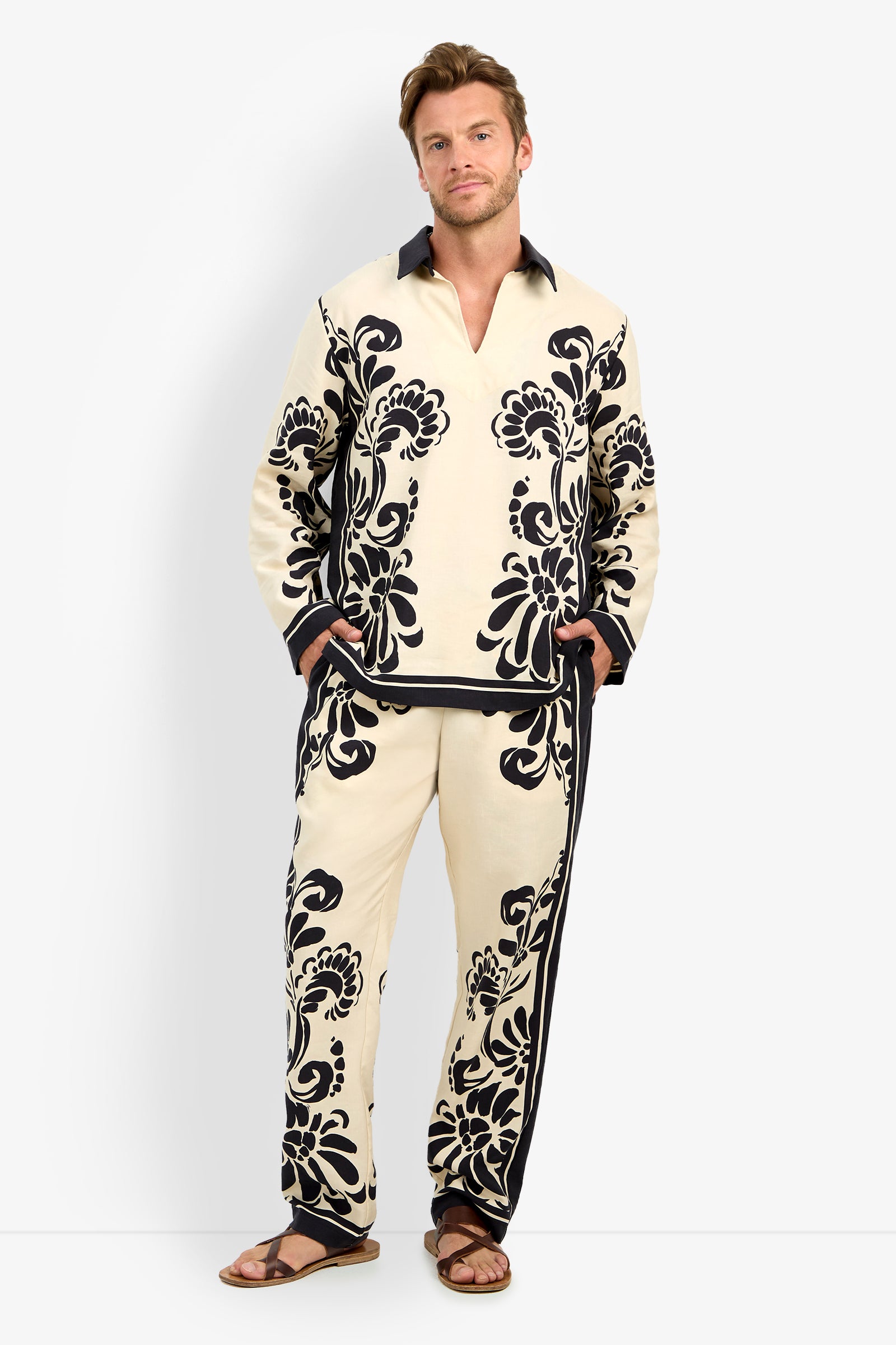 A man stands against a white background wearing the HOLMES PULLOVER in Tivoli, featuring bold black floral patterns and a matching set, paired with brown sandals and his hands in his pockets.