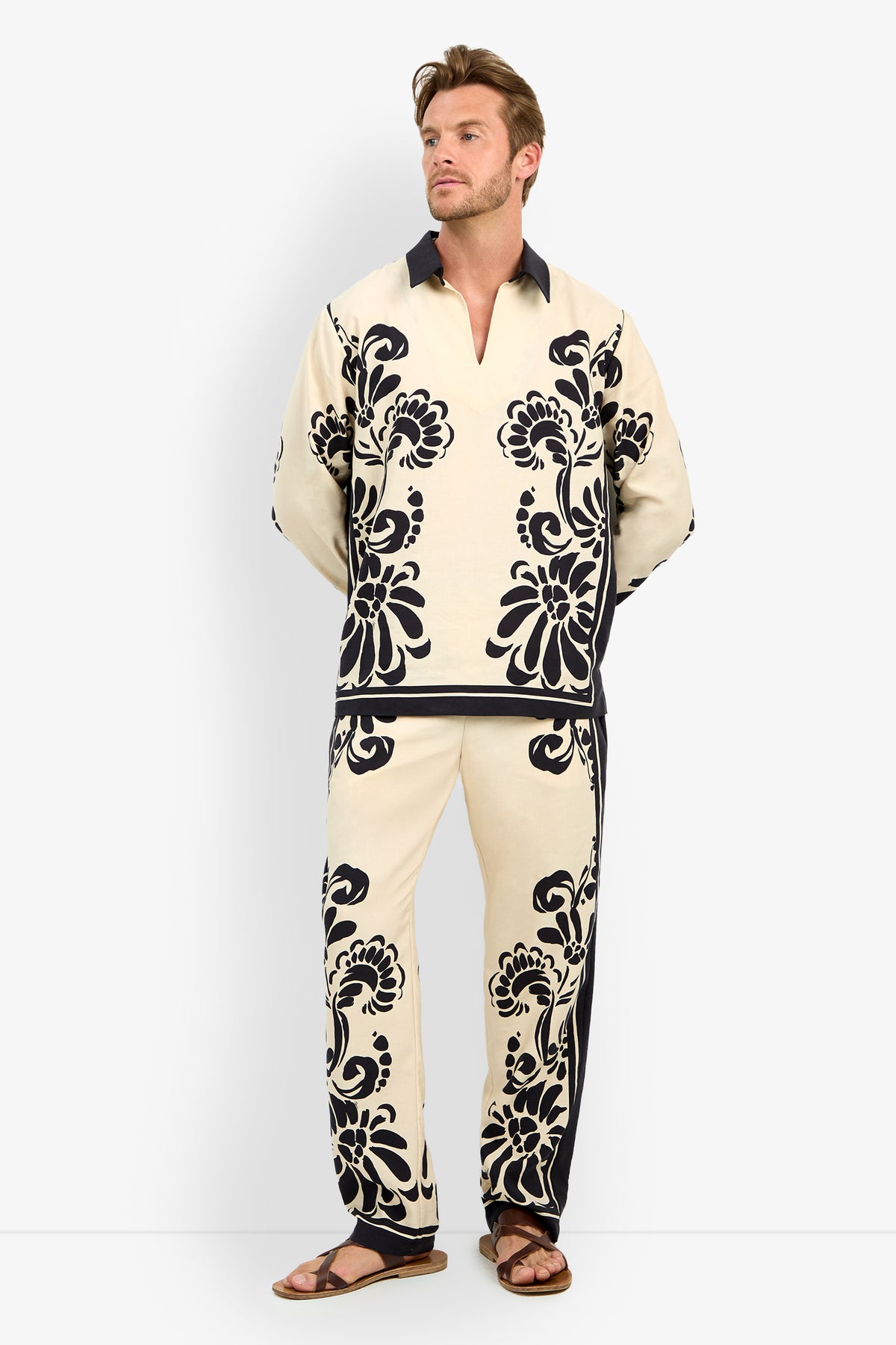 A man with light brown hair and a short beard wears the HOLMES PULLOVER, a cream-colored shirt featuring bold black floral patterns, paired with matching pants and brown sandals, standing against a plain white background.