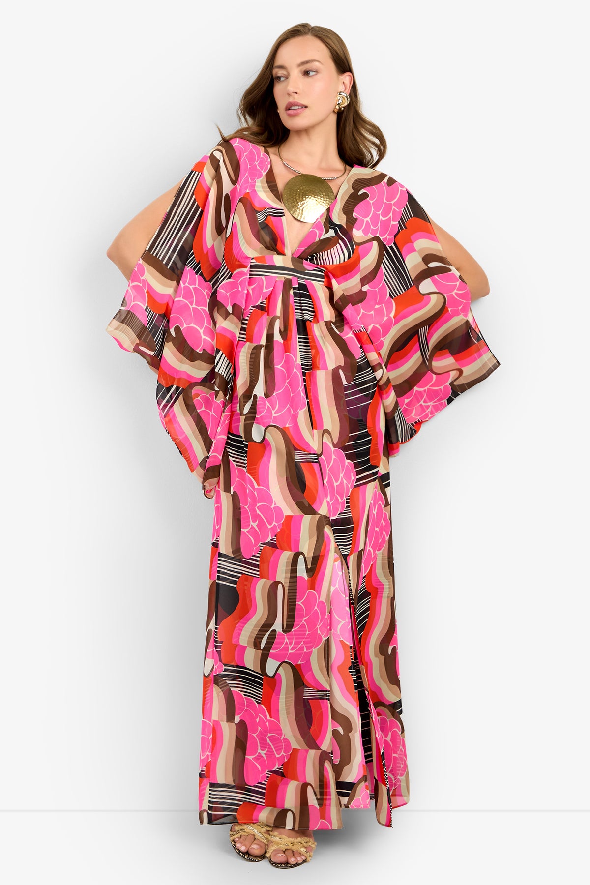 A woman wears the BLOSSOM DRESS, a flowing, long-sleeve maxi with a bold abstract geometric print in pink, brown, orange, and white. She pairs it with gold earrings and sandals against a plain white background.