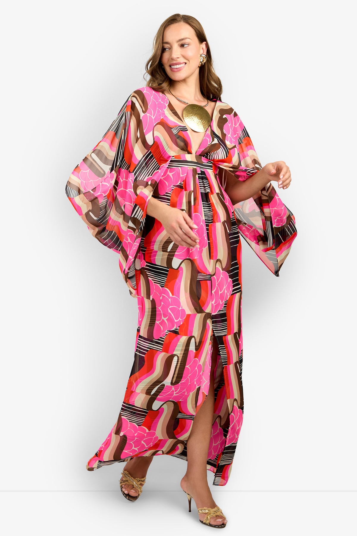 A woman smiles in the BLOSSOM DRESS, a colorful maxi with bold pink, red, black, and beige patterns, wide sleeves, and a front slit. She accessorizes with a gold necklace, earrings, and open-toe heels.