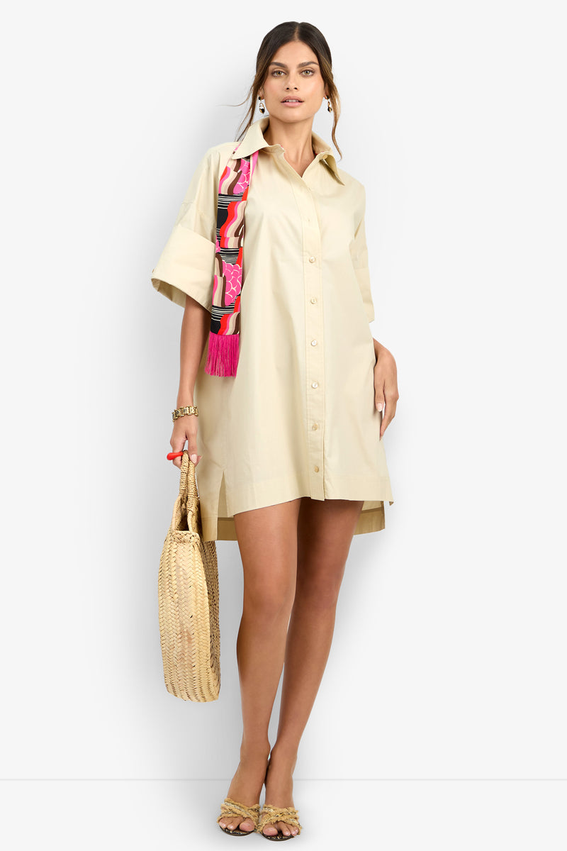 A woman wears the OPPONENT DRESS, a beige button-up shirt dress with rolled sleeves. She accessorizes with a woven straw bag, open-toe sandals, gold jewelry, and drapes a colorful patterned scarf over one shoulder.