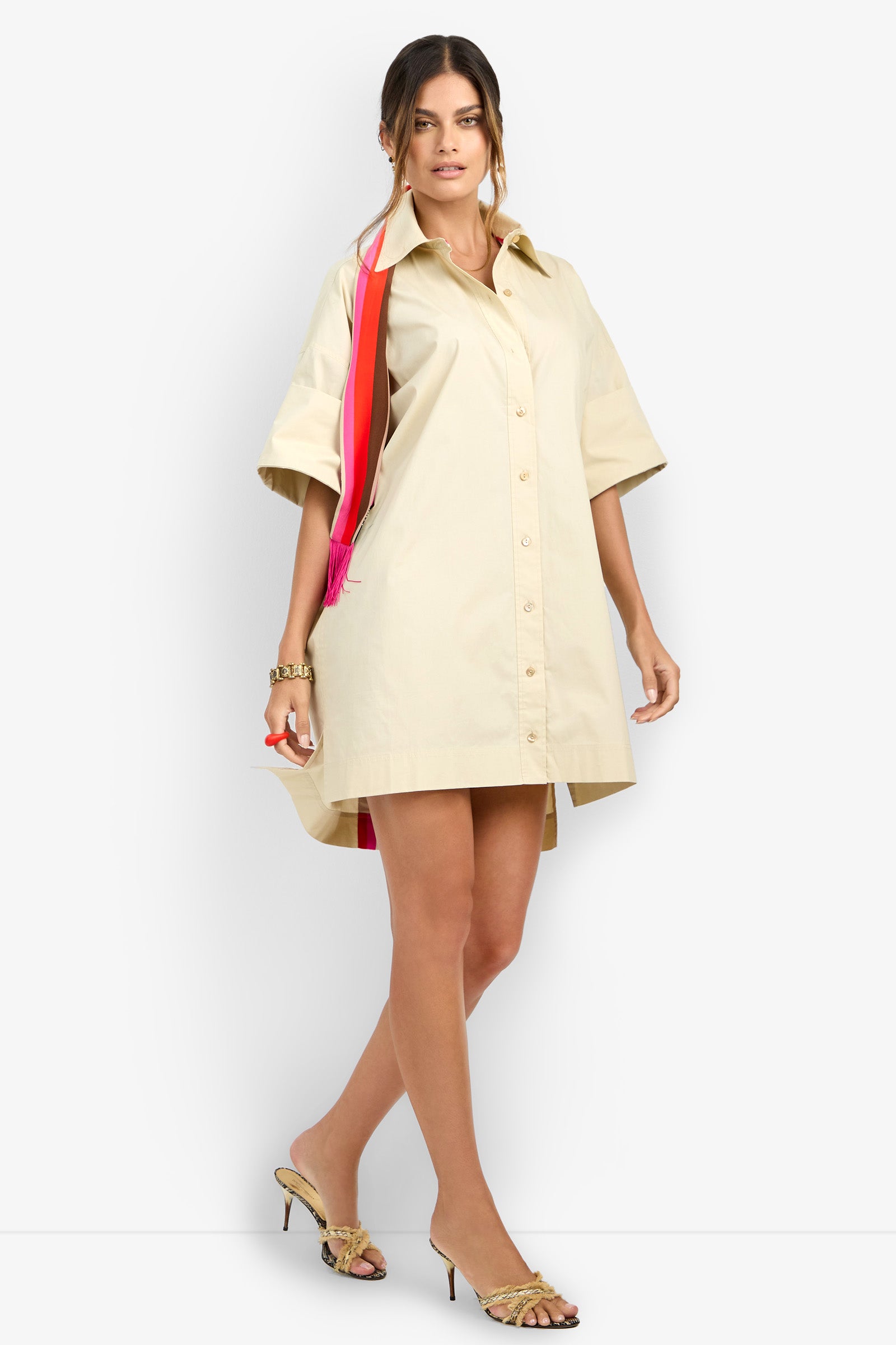 A woman poses against a plain white background wearing the OPPONENT DRESS, a loose beige button-up shirt dress with short sleeves, styled with gold bracelets, heeled sandals, and a red and pink shoulder bag.