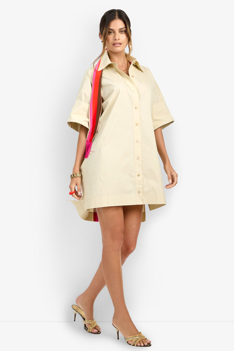 A woman poses against a plain white background wearing the OPPONENT DRESS, a loose beige button-up shirt dress with short sleeves, styled with gold bracelets, heeled sandals, and a red and pink shoulder bag.